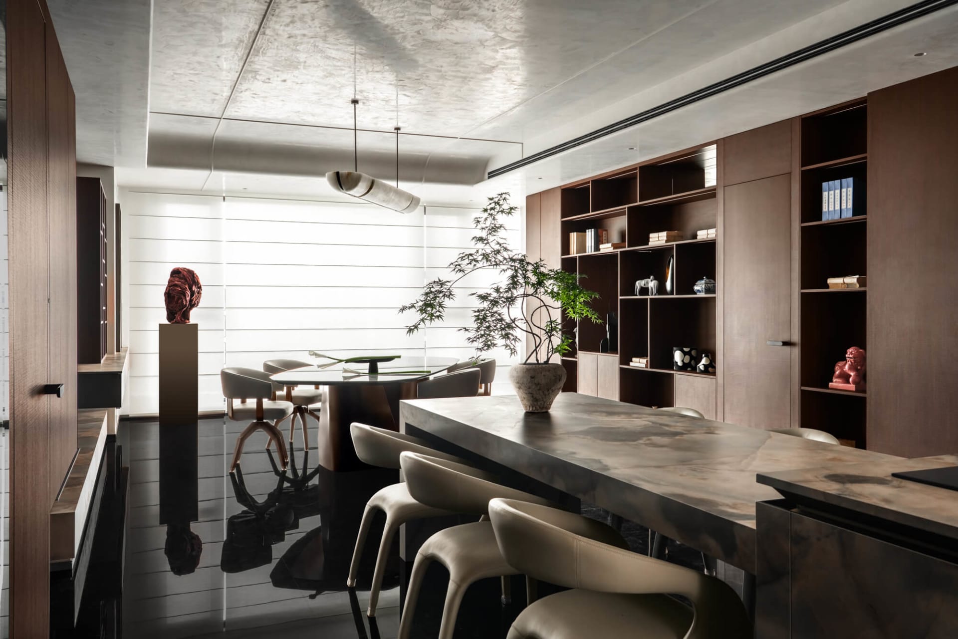 Modern luxury dining room featuring a long marble island with beige leather chairs, a round glass dining table, built-in walnut bookshelves, and a large potted tree against a white slatted wall.