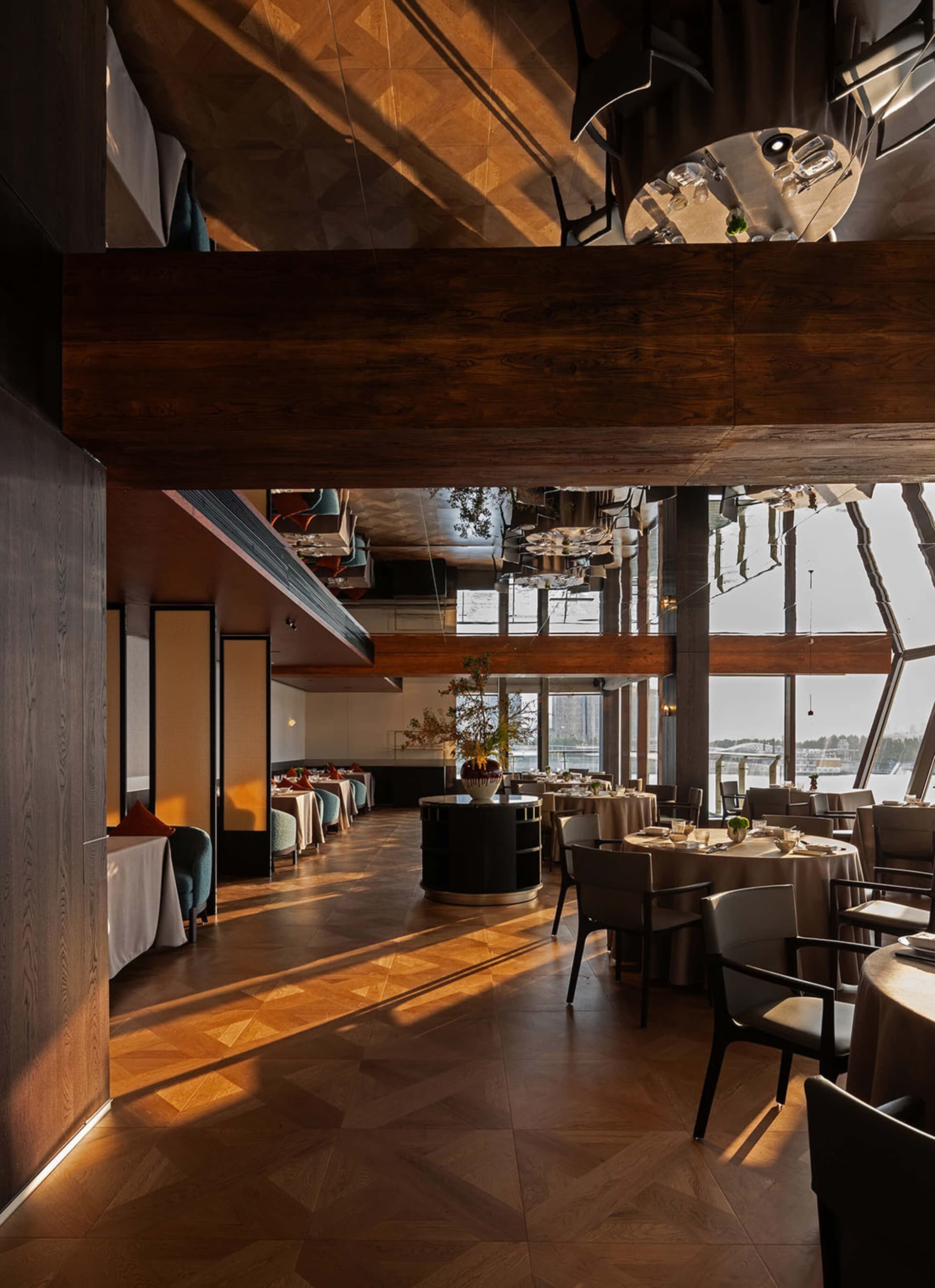 Luxurious multi-level restaurant interior featuring dark wood ceilings, herringbone parquet flooring, round dining tables with neutral linens, and large windows overlooking a waterfront view.