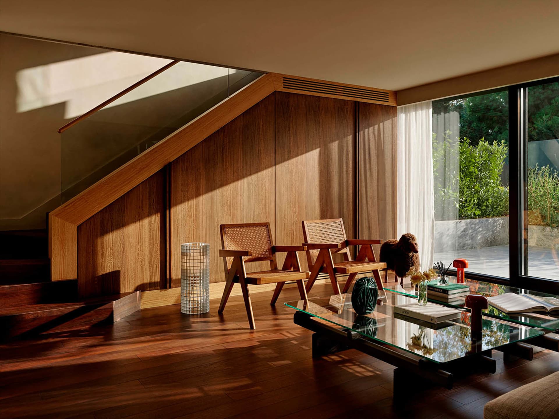 Modern living room featuring warm teak wood paneling, two wooden armchairs with cane backs, a glass coffee table, and dramatic sunlight streaming from a high clerestory window under a staircase.