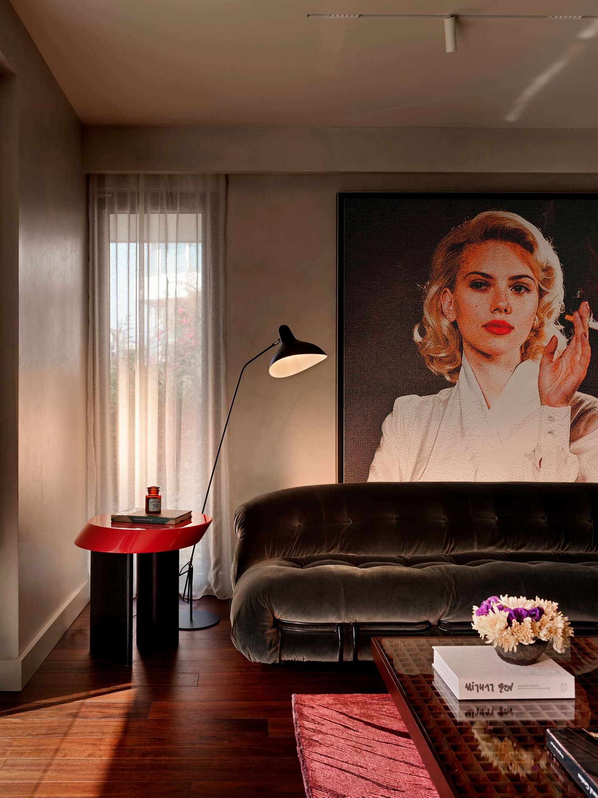 Modern living room with a dark green velvet tufted sofa, glossy red side table, black floor lamp, and a large framed portrait of Scarlett Johansson on a neutral wall with sheer curtains.