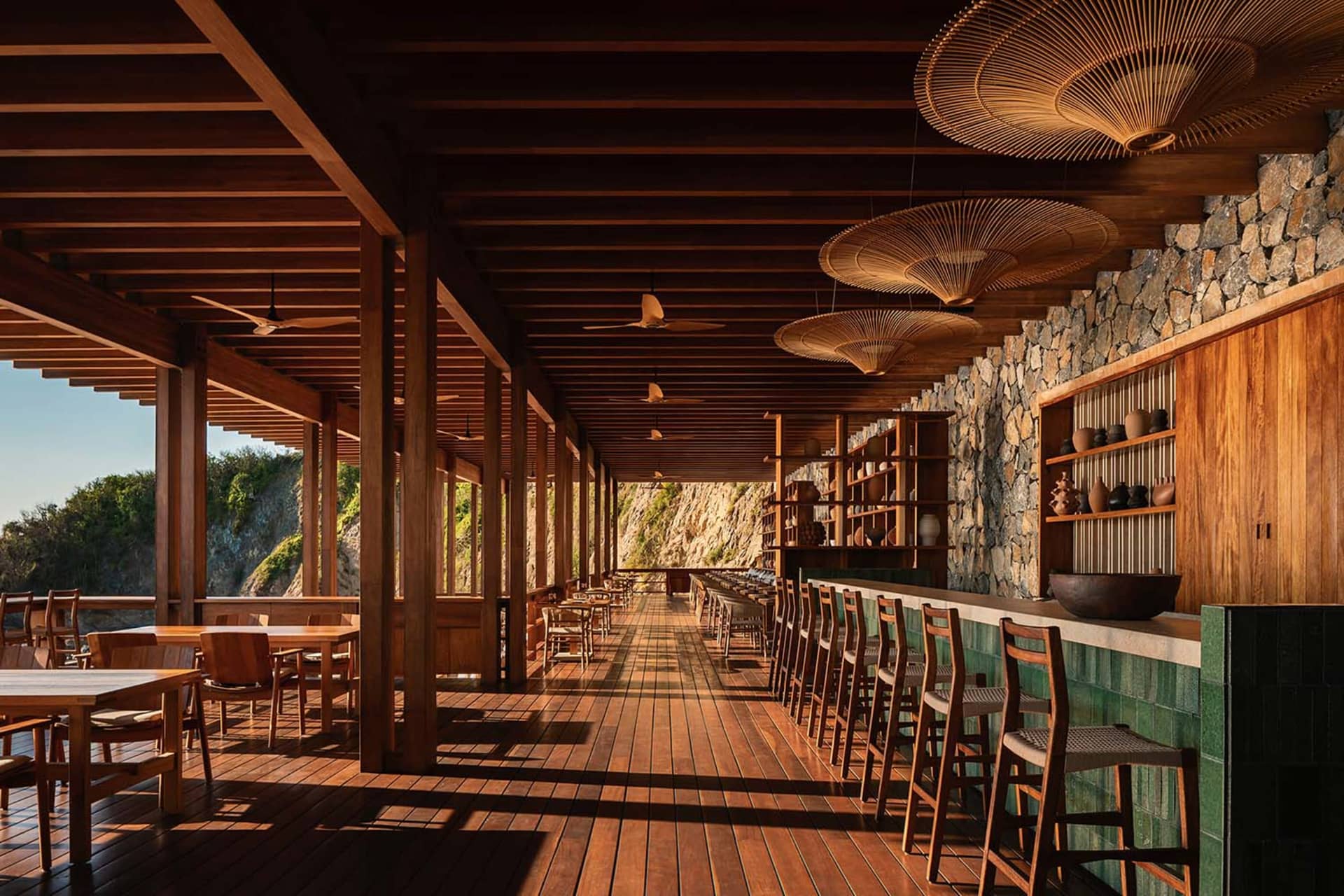 Long open-air wooden veranda with teak flooring and ceiling, featuring a stone wall, green tiled bar counter, woven rattan pendant lamps, and rows of wooden tables and chairs overlooking a dramatic coastal cliff.