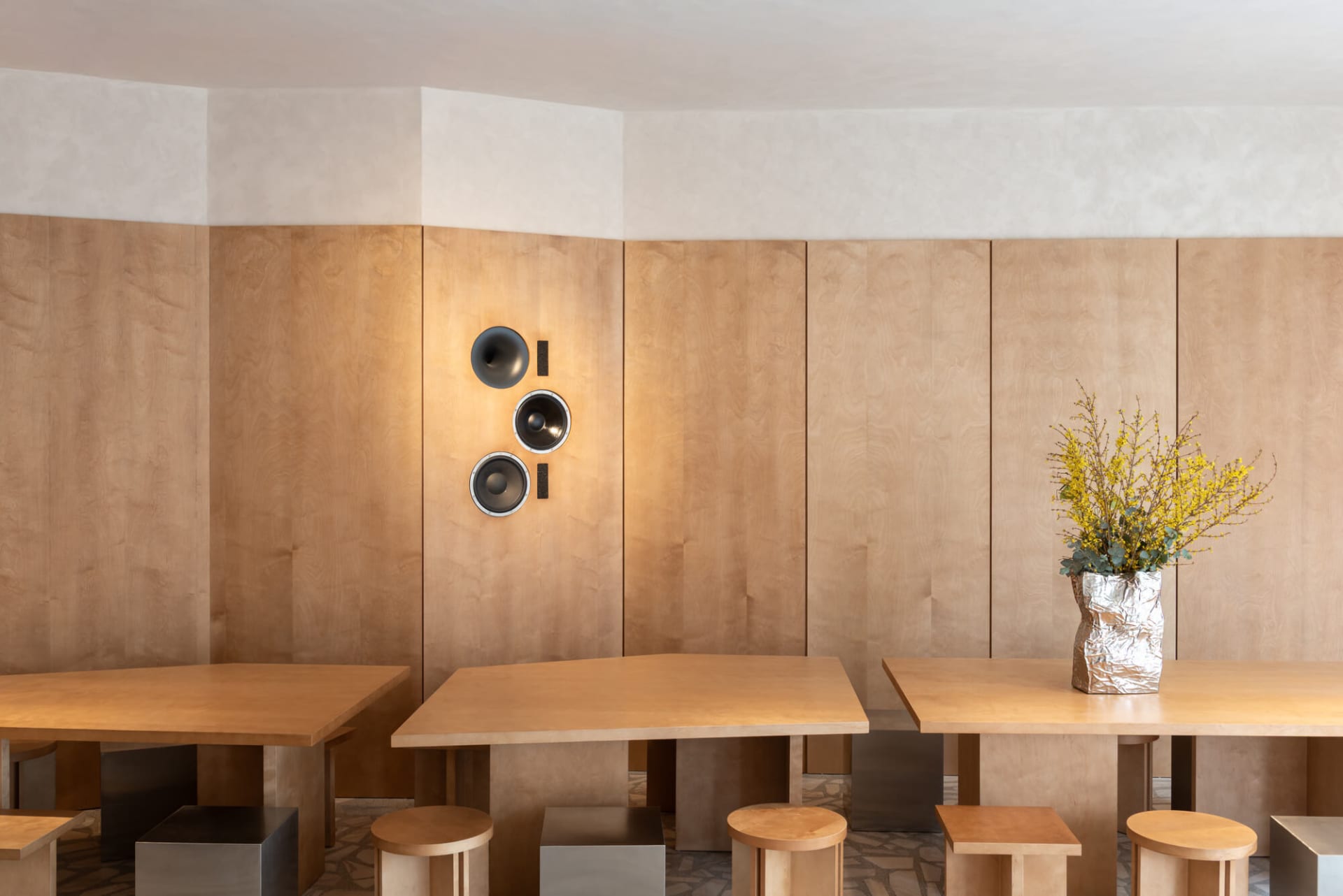Modern minimalist dining room featuring light oak wood paneling, three sculptural black and silver wall speakers, solid oak rectangular tables, and mixed metal and wood stools, with a vase of yellow forsythia branches.