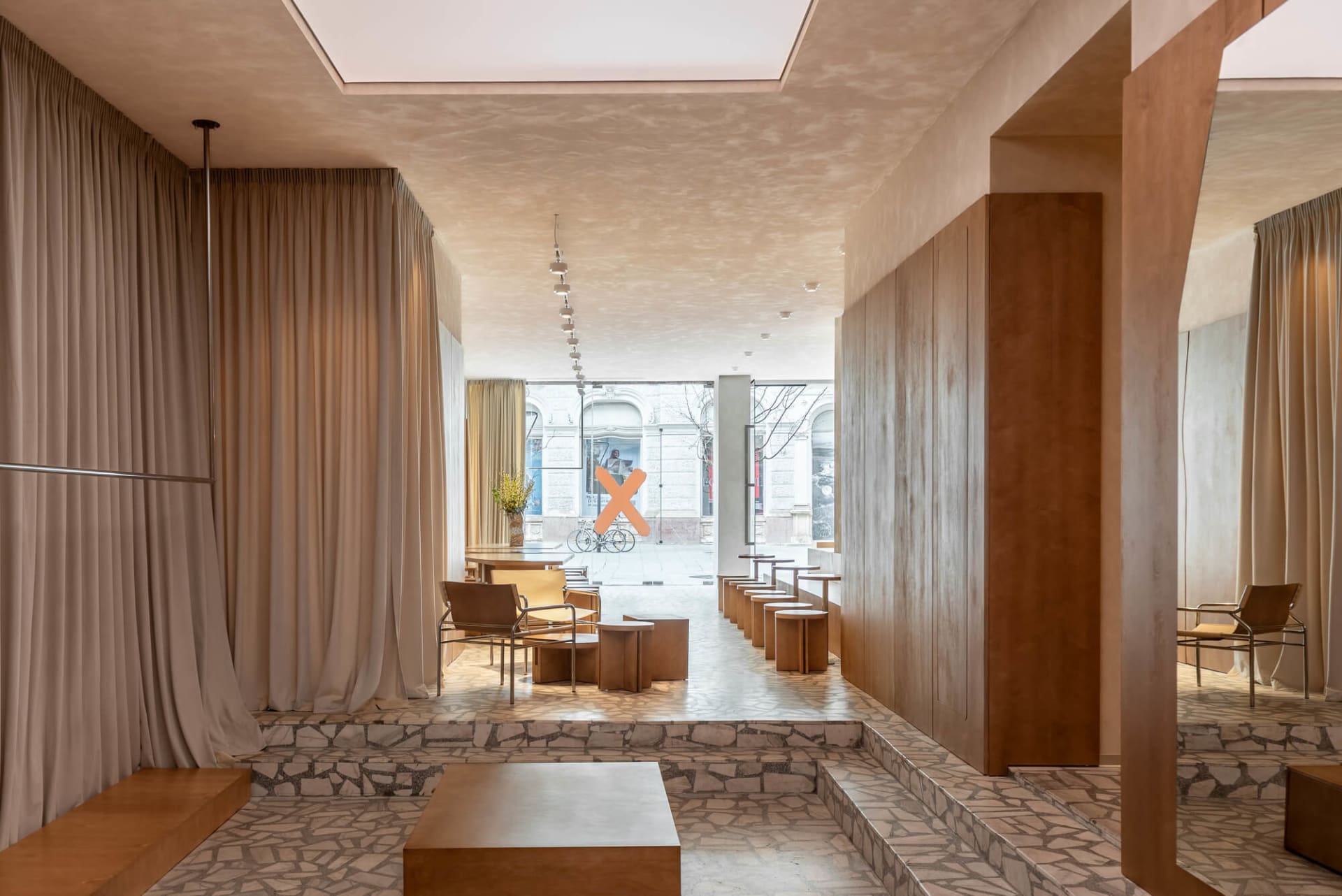 Modern minimalist boutique interior featuring irregular terrazzo flooring and steps, warm oak wood paneling, neutral linen curtains, and contemporary seating areas with wooden tables and leather chairs.