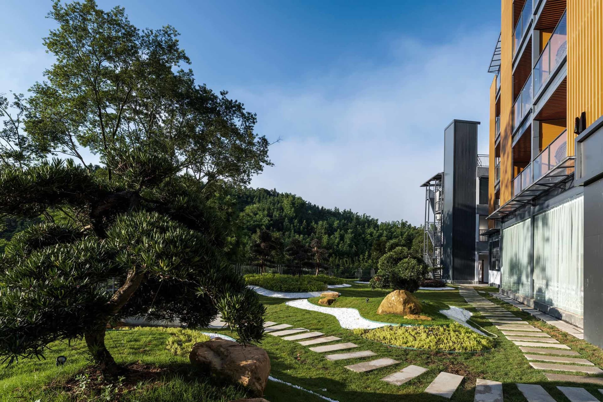 Lush Japanese-inspired garden featuring a large pine tree, winding white gravel paths, large natural boulders, stepping stone pathways, and a modern building with yellow wooden cladding on the right.