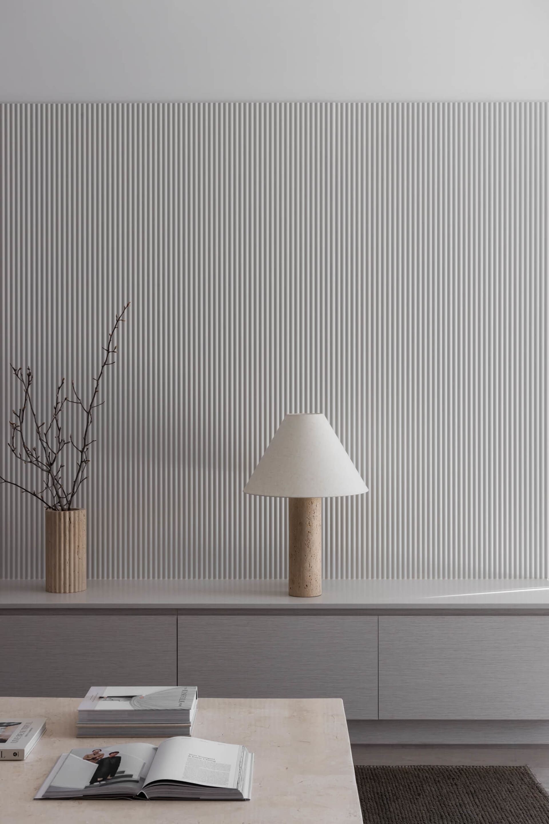 Modern minimalist living room featuring a white fluted wall, grey floating console, travertine table lamp with fabric shade, and a travertine coffee table with open books and a wooden vase with dried branches.