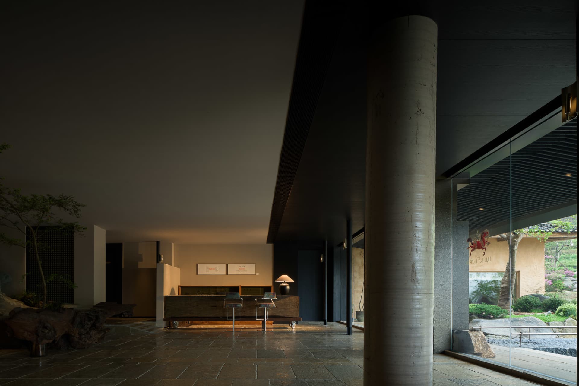 Modern wabi-sabi style hotel lobby featuring a massive concrete pillar, dark stone tile floor, reclaimed wood reception desk, and large glass windows overlooking a Japanese garden.
