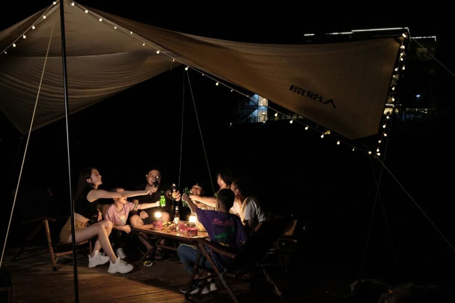 A group of adults and children sitting around a wooden table under a large beige canvas camping tent at night, illuminated by warm string lights, toasting with drinks.