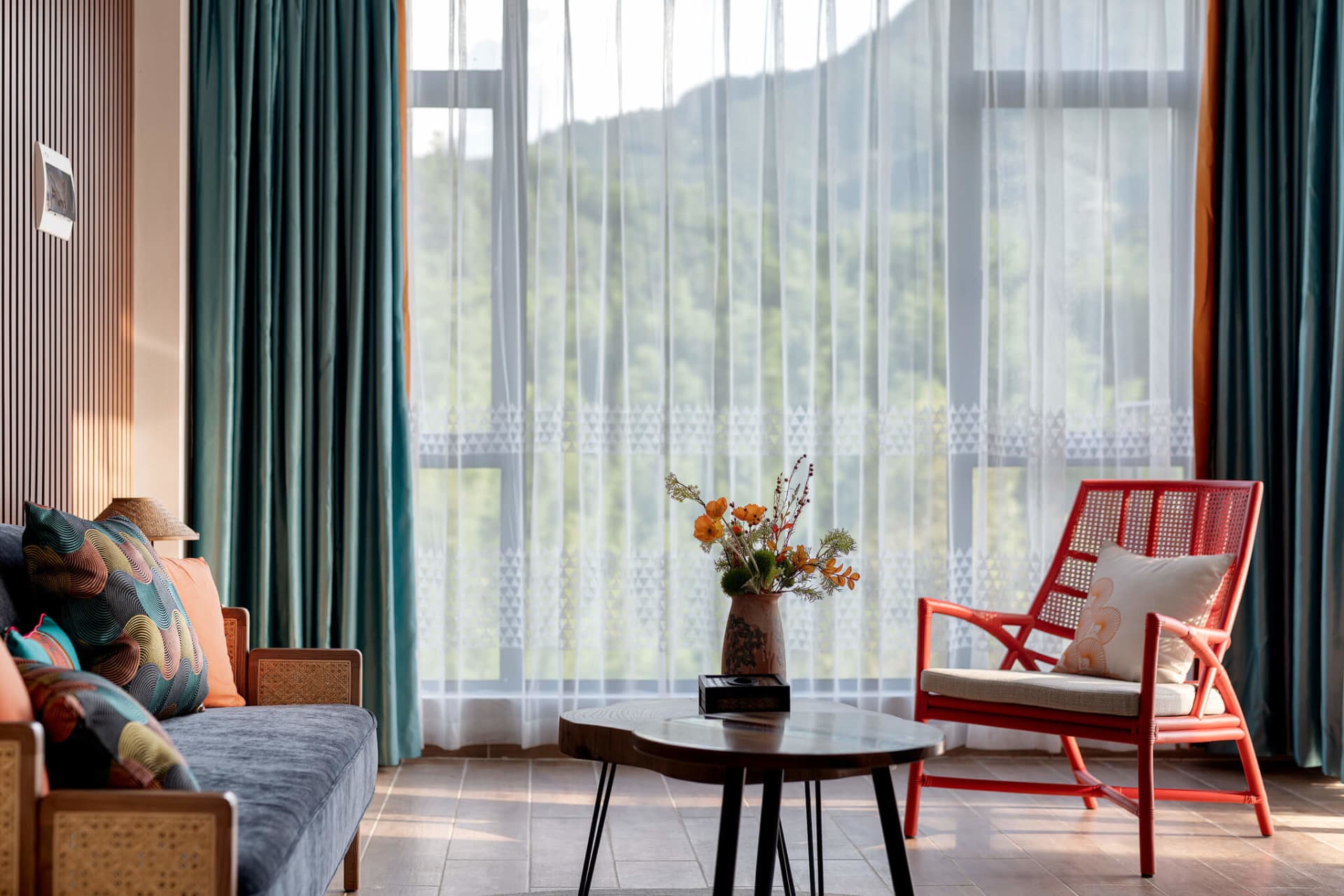Modern living room featuring a blue sofa with patterned cushions, a bright red rattan armchair, wooden coffee table with flower vase, and large windows overlooking green mountains.