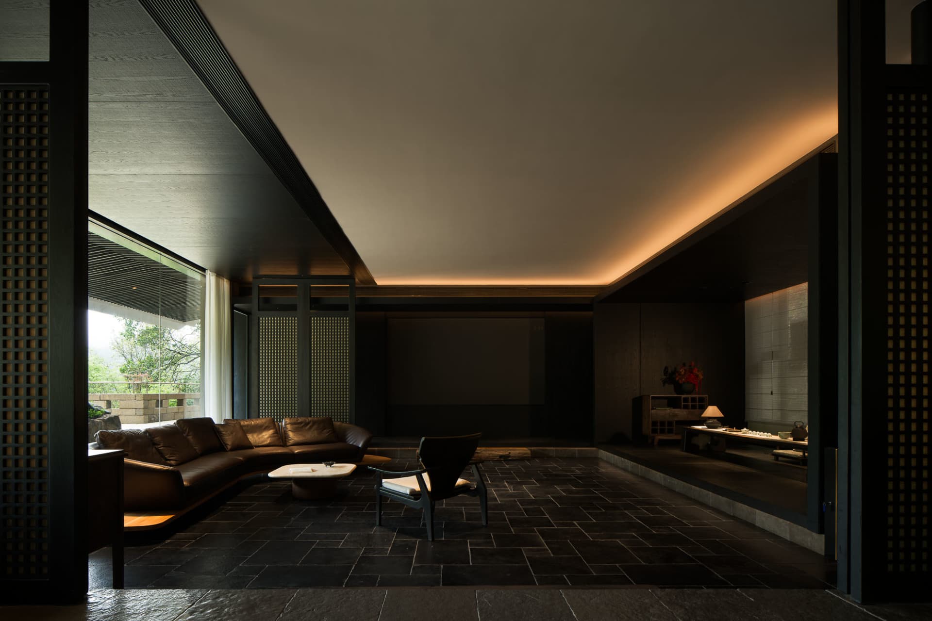 Modern dark-toned living room featuring a curved brown leather sofa, single black armchair, large projection screen, and large glass doors opening to a mountain view.