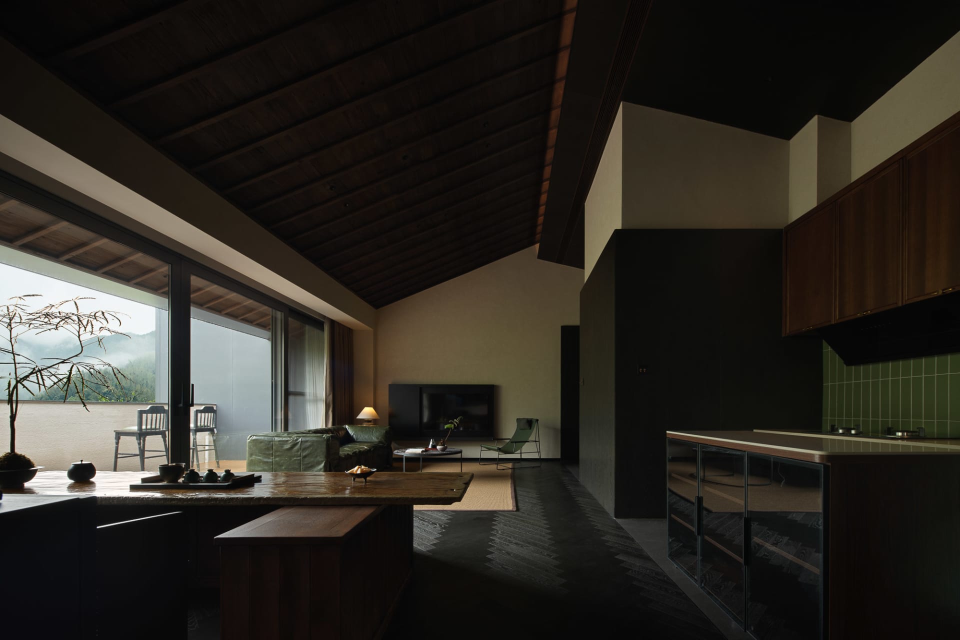 Modern dark wood interior of a living room and kitchen with high pitched timber ceiling, large glass doors opening to a mountain view, green leather sofa, and minimalist furniture.