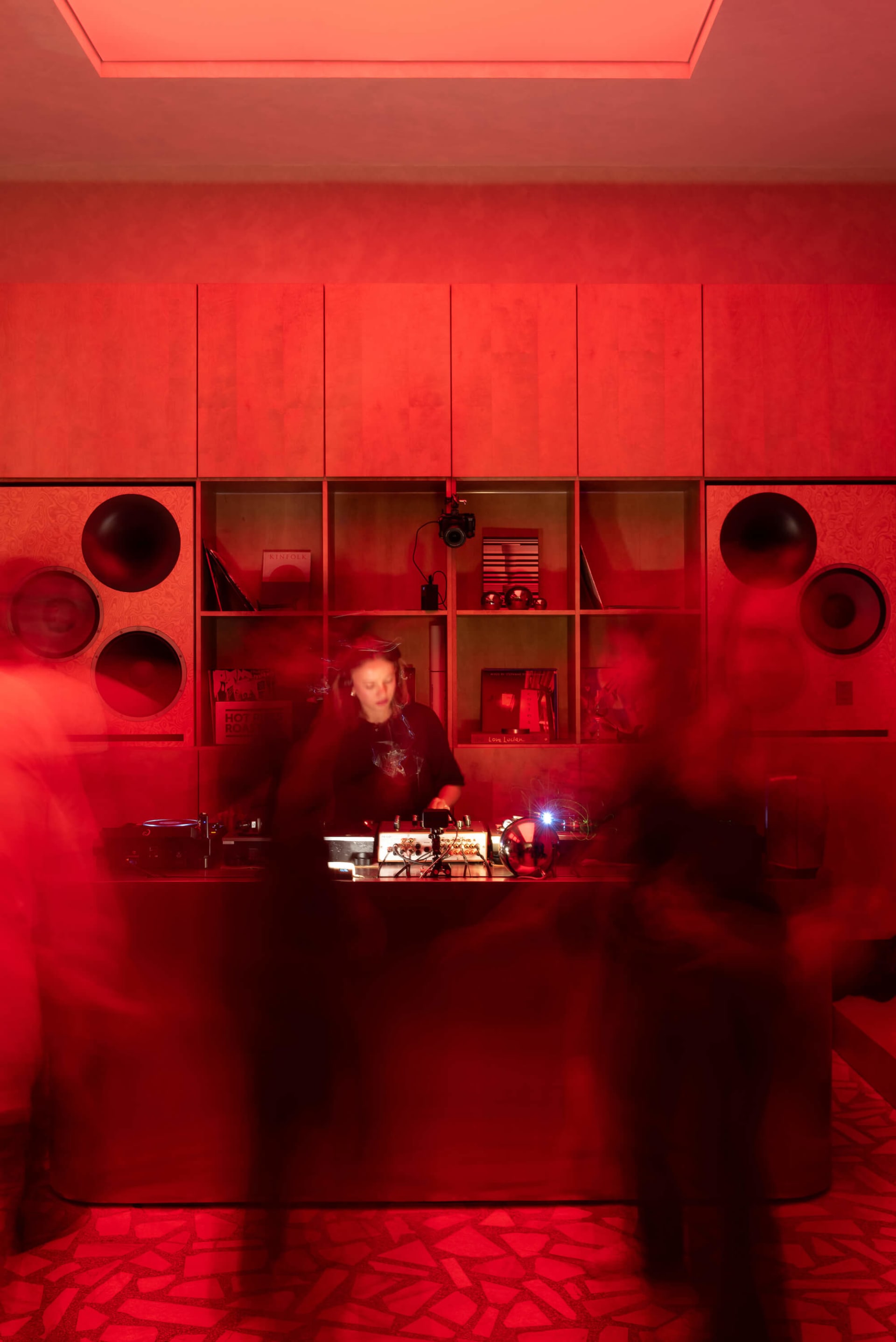 A female DJ performing at a wooden booth surrounded by large speakers and red-lit cabinetry, with motion-blurred dancers in the foreground creating a dynamic nightclub atmosphere.