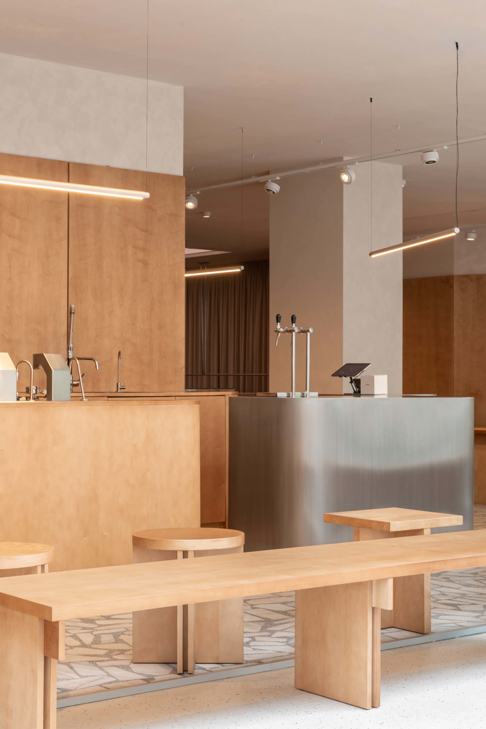 Modern minimalist café interior featuring a long birch plywood communal table, round stools, stainless steel service counter with beer taps, and linear pendant lighting.