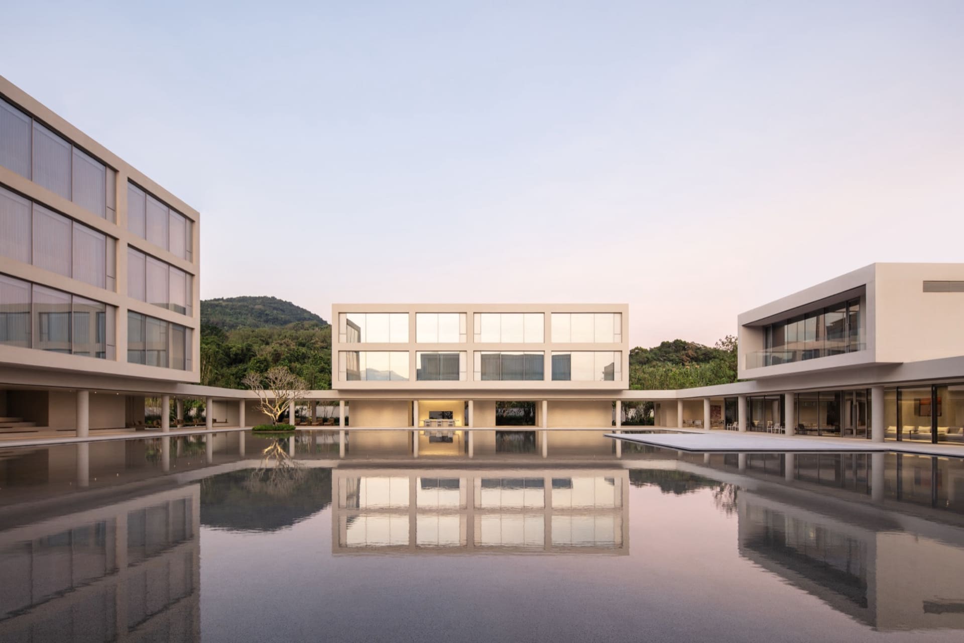 Modern minimalist courtyard house with large reflecting pool at dawn, surrounded by lush green hills.