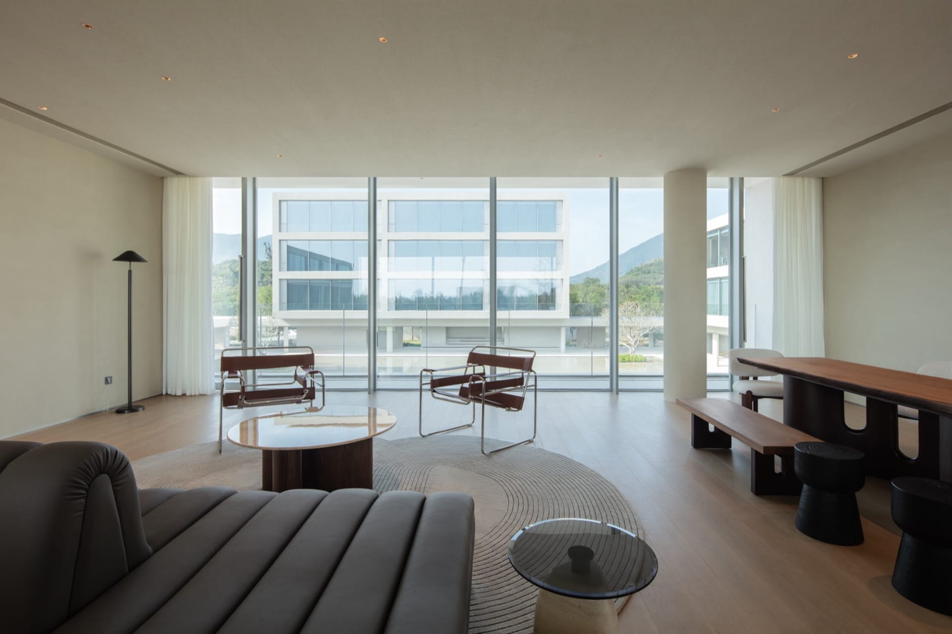 Modern minimalist living room featuring brown leather and chrome lounge chairs, walnut coffee tables, a curved rug, and large floor-to-ceiling windows overlooking mountains and contemporary buildings.