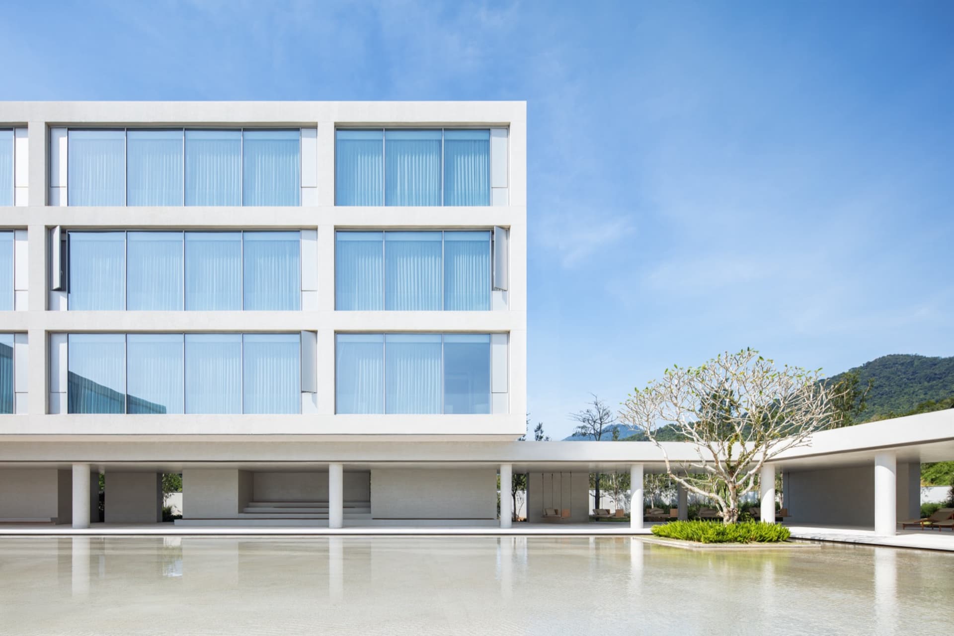 Modern minimalist concrete building with large glass windows, elevated on pillars, overlooking a calm reflecting pool with a single tree and mountain view in the background.