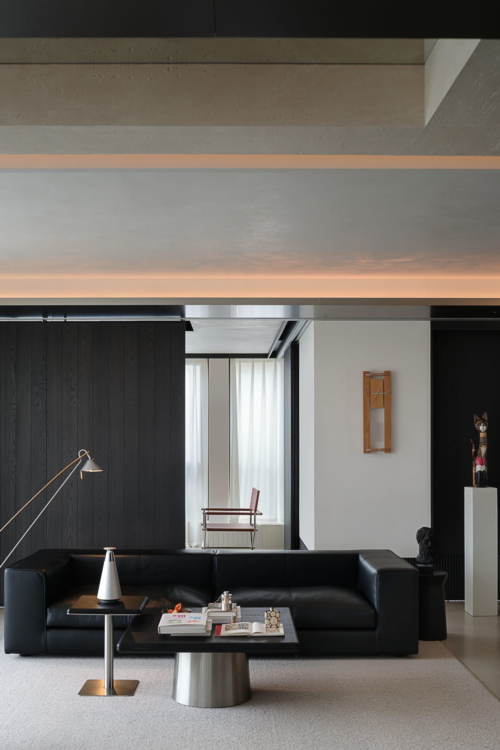 Modern minimalist living room featuring exposed concrete ceiling, black leather sofa, dark wood wall paneling, and sculptural lighting.