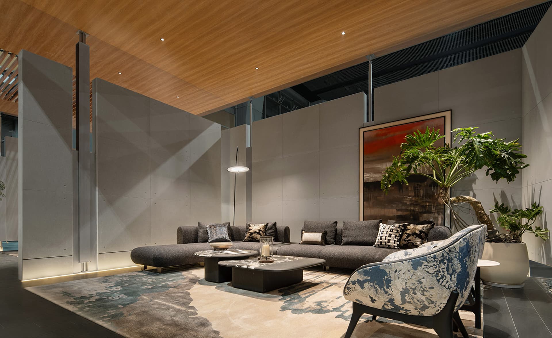 Modern living room with grey L-shaped sectional sofa, blue and white patterned accent chair, concrete walls, wooden ceiling, and large potted plant.