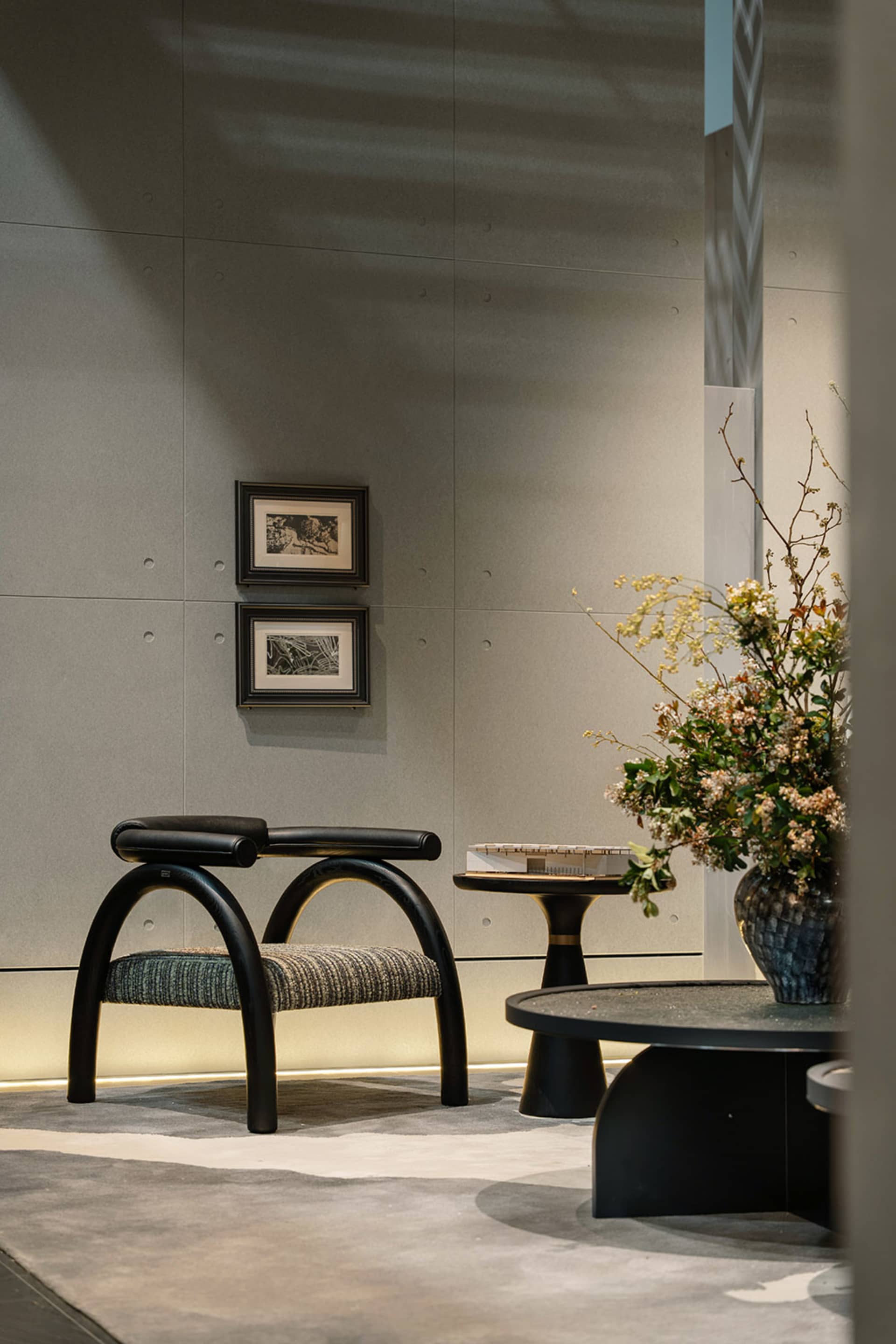 Modern minimalist entryway featuring a black arched accent chair with textured seat, round black side table, large floral arrangement in a blue ceramic vase, and two framed artworks on a grey concrete wall.