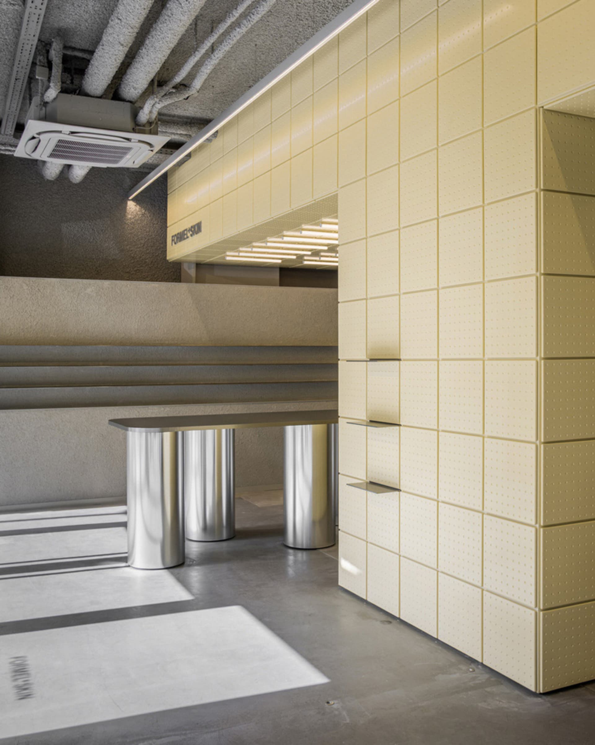 Modern commercial interior with a pale yellow perforated metal wall, integrated floating shelves, cylindrical stainless steel bench, exposed concrete ceiling with visible ductwork and air conditioning unit.