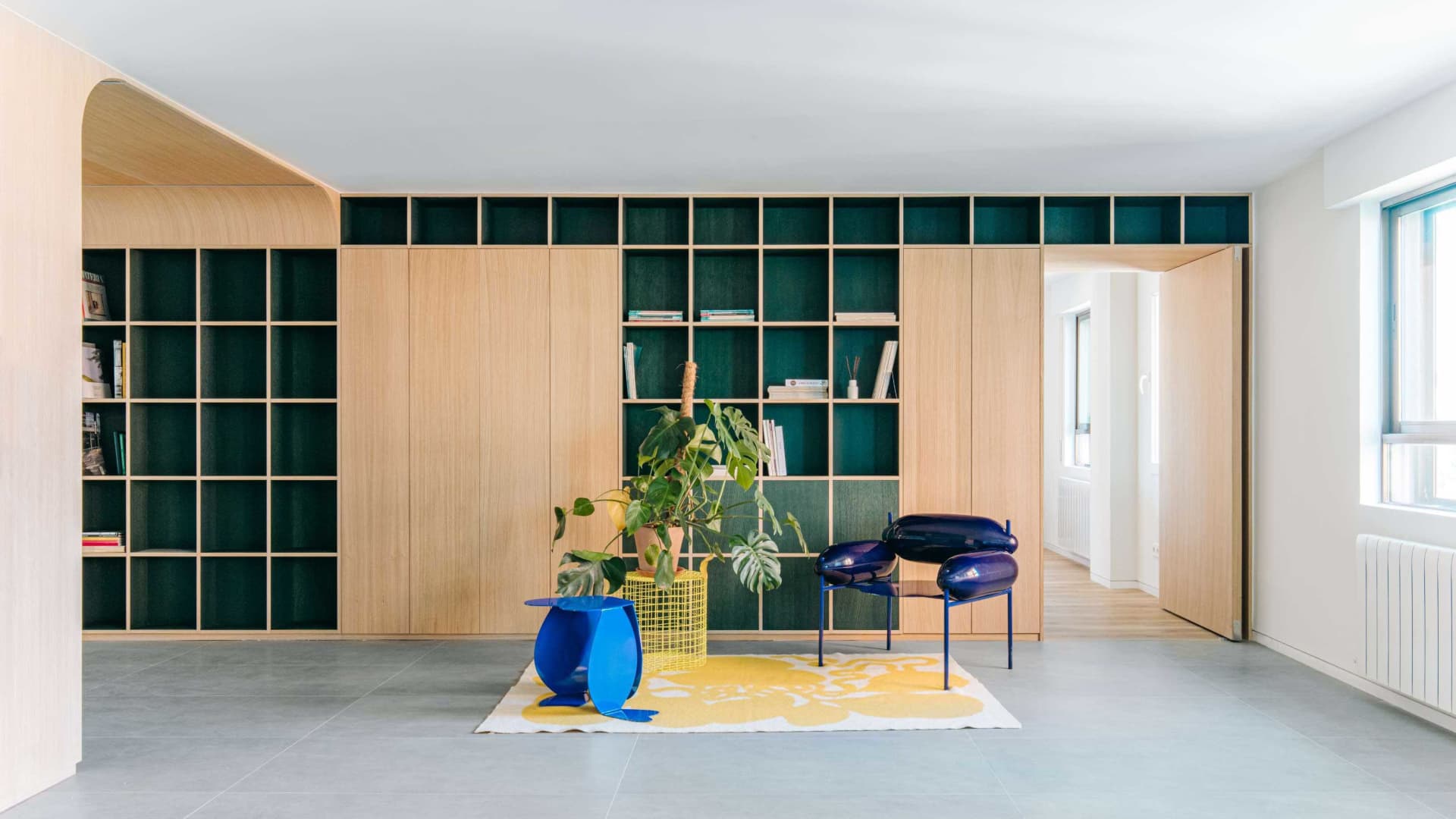 Modern living room featuring a large oak built-in shelving unit with dark green backs, bright blue sculptural chairs, a yellow and white rug, and a large monstera plant.