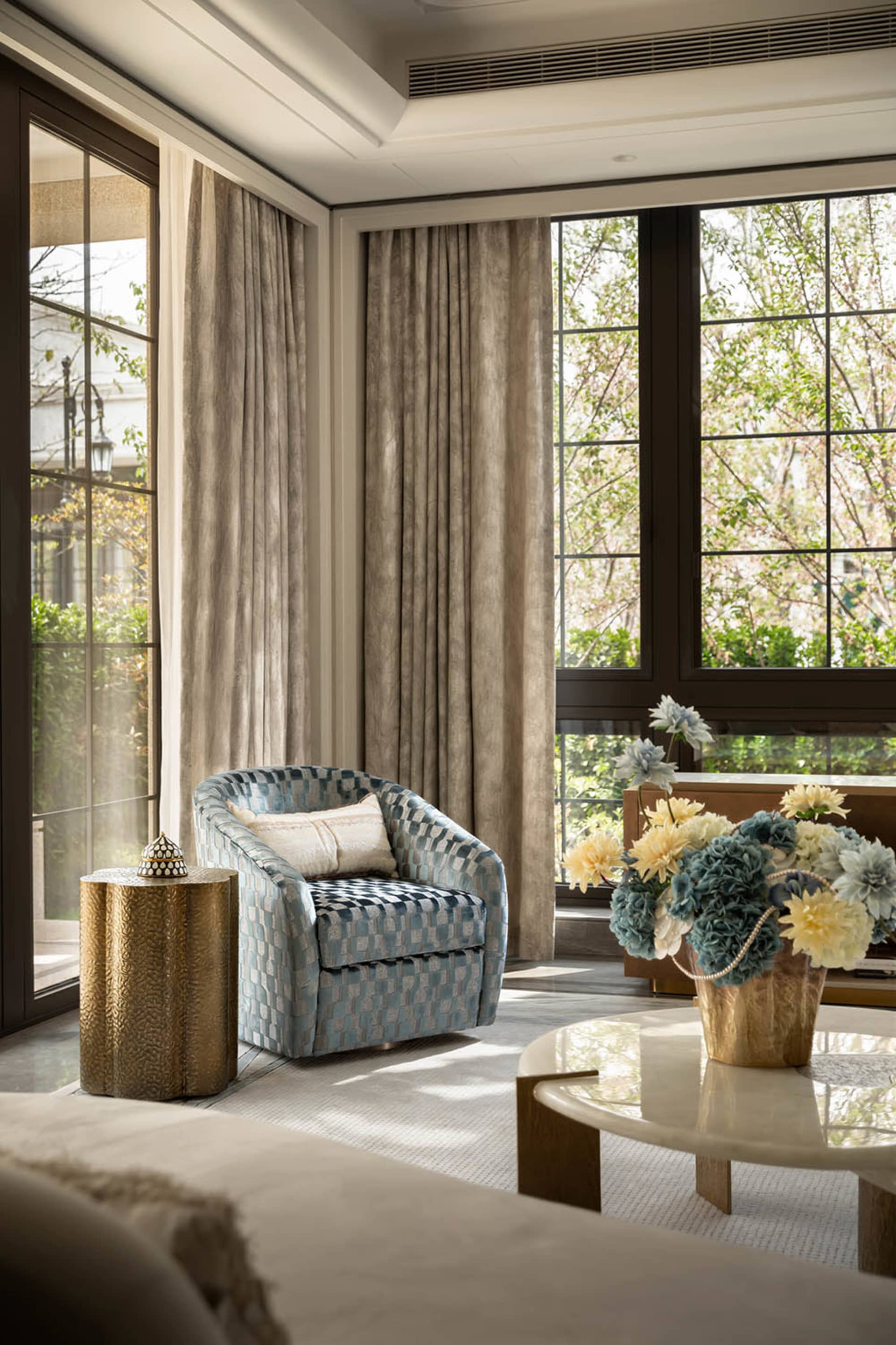A contemporary living room corner featuring a blue and white checkered upholstered armchair, a textured gold cylindrical side table, a large marble coffee table with a floral arrangement in a brass vase, and floor-to-ceiling windows with neutral curtains.