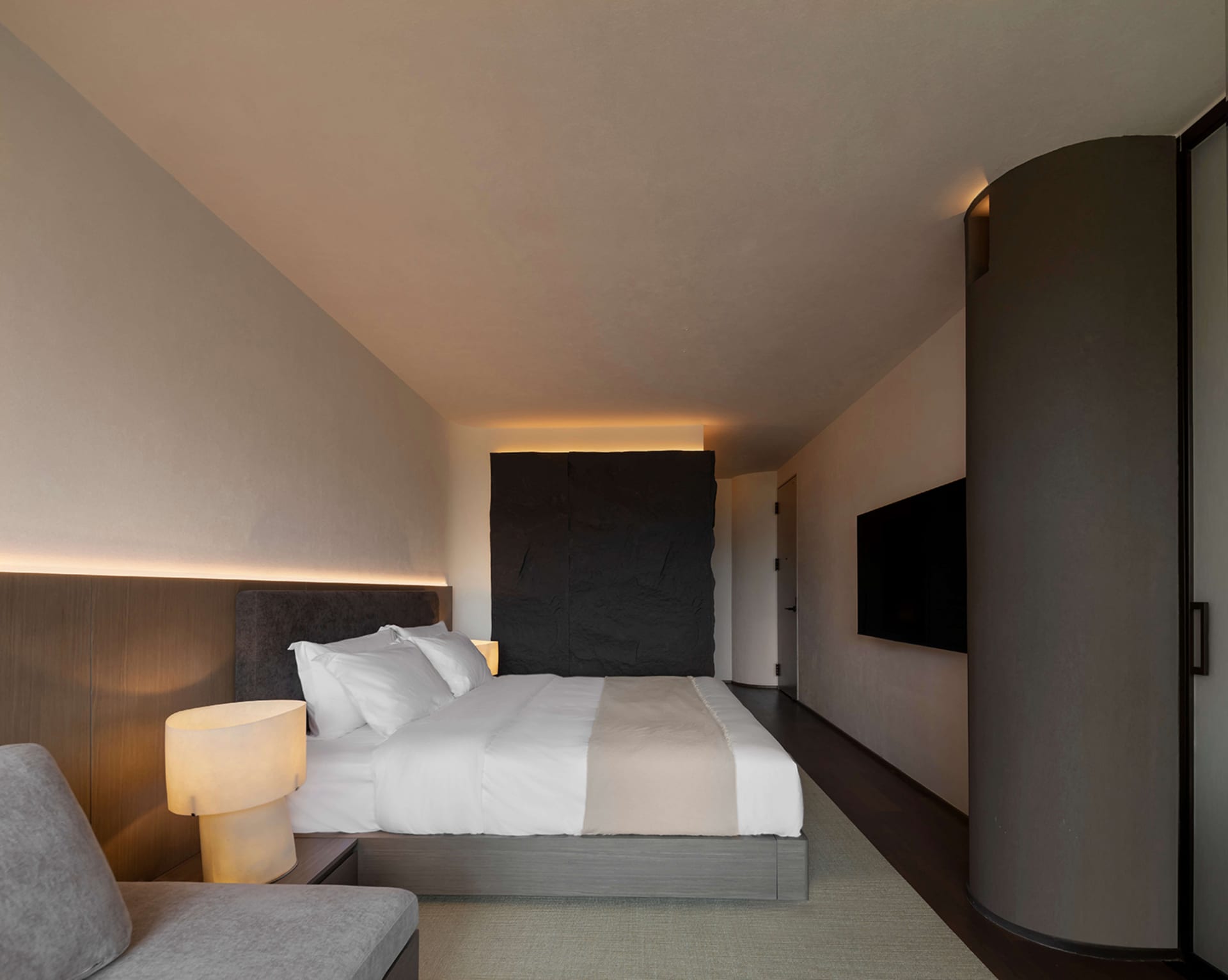 Modern minimalist bedroom interior with a large dark stone feature wall behind the bed, warm LED cove lighting, neutral bedding, and a curved dark wall panel on the right.