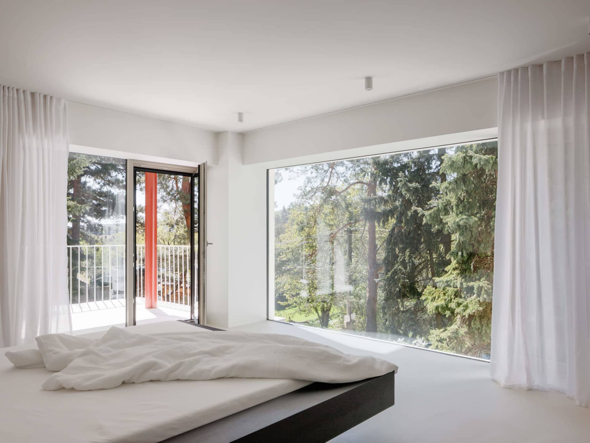 Minimalist bedroom with low platform bed dressed in white linen, sheer white curtains, and large windows overlooking a lush green forest.