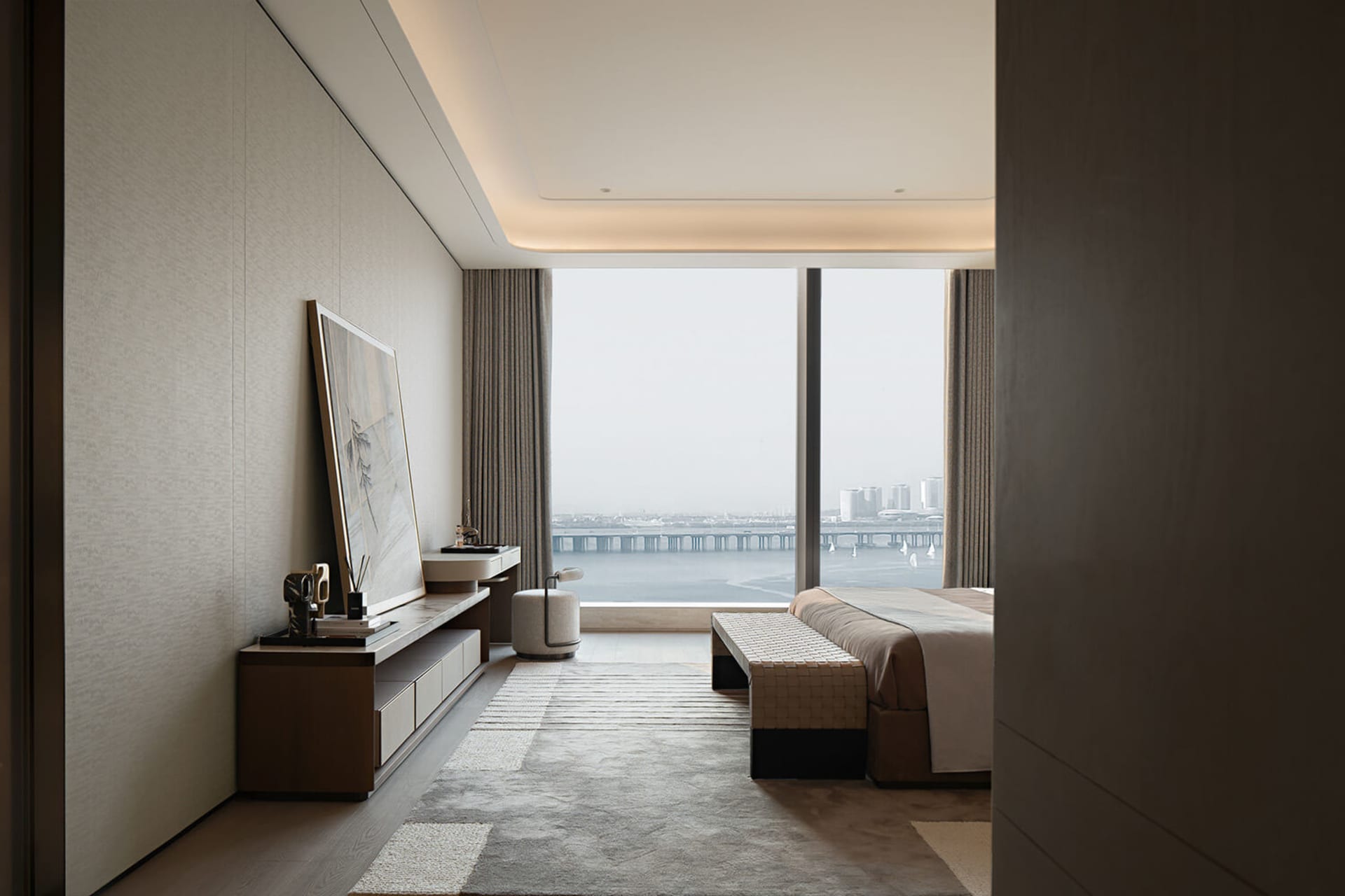 Modern luxury bedroom interior featuring a large harbor view window, walnut media console, textured area rug, and woven bench at the foot of the bed.