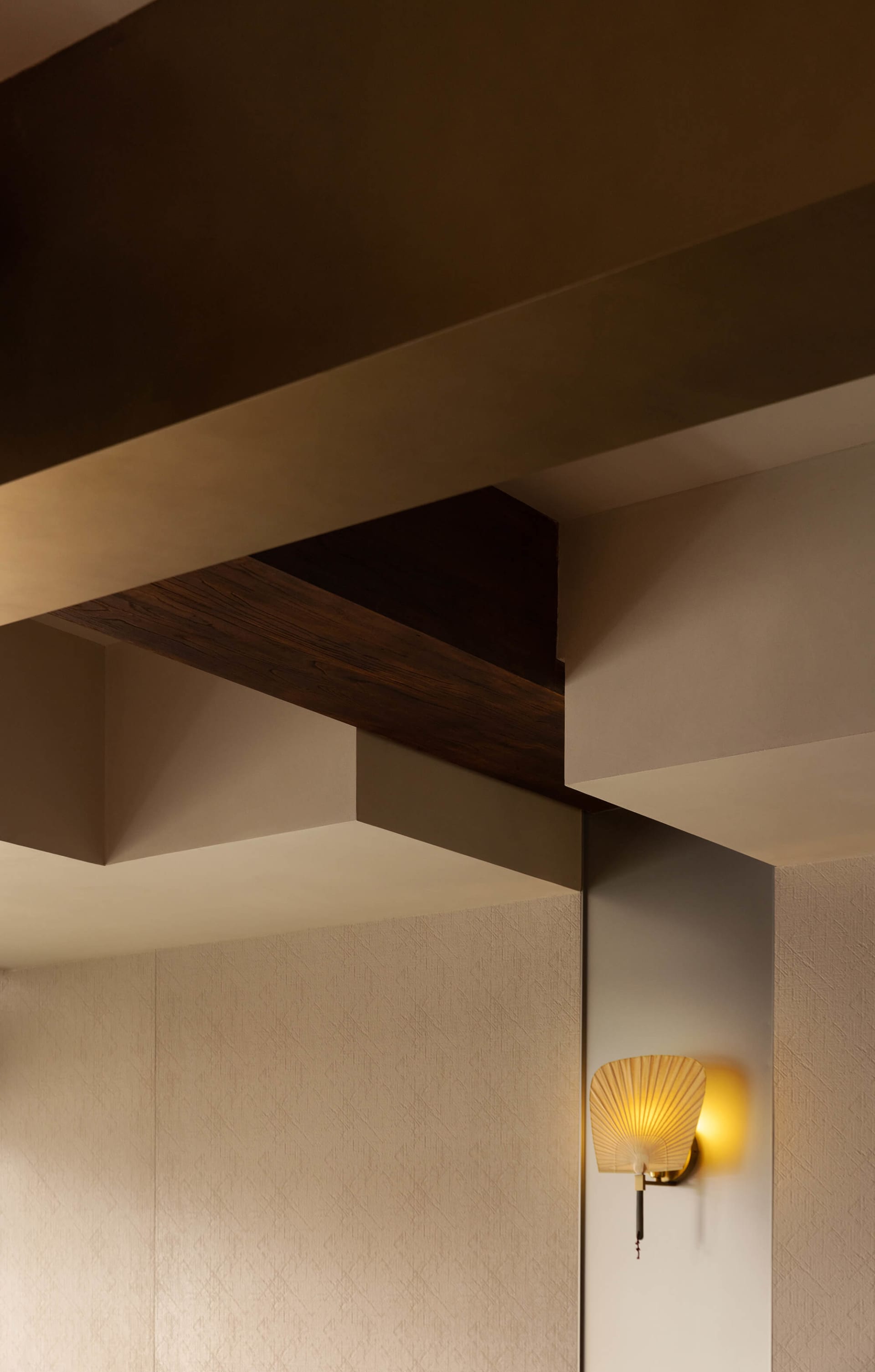 Close-up view of a modern ceiling featuring geometric beige planes, a dark walnut wood beam, and a lit golden fan-shaped wall sconce on a textured wall.