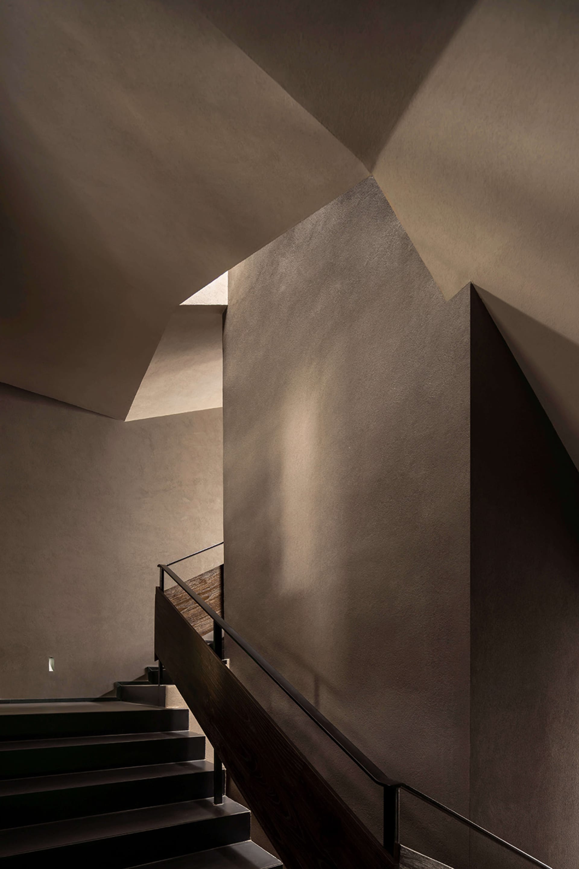 Modern concrete staircase with dark wood handrail ascending under angular geometric ceiling planes that create strong directional shadows.