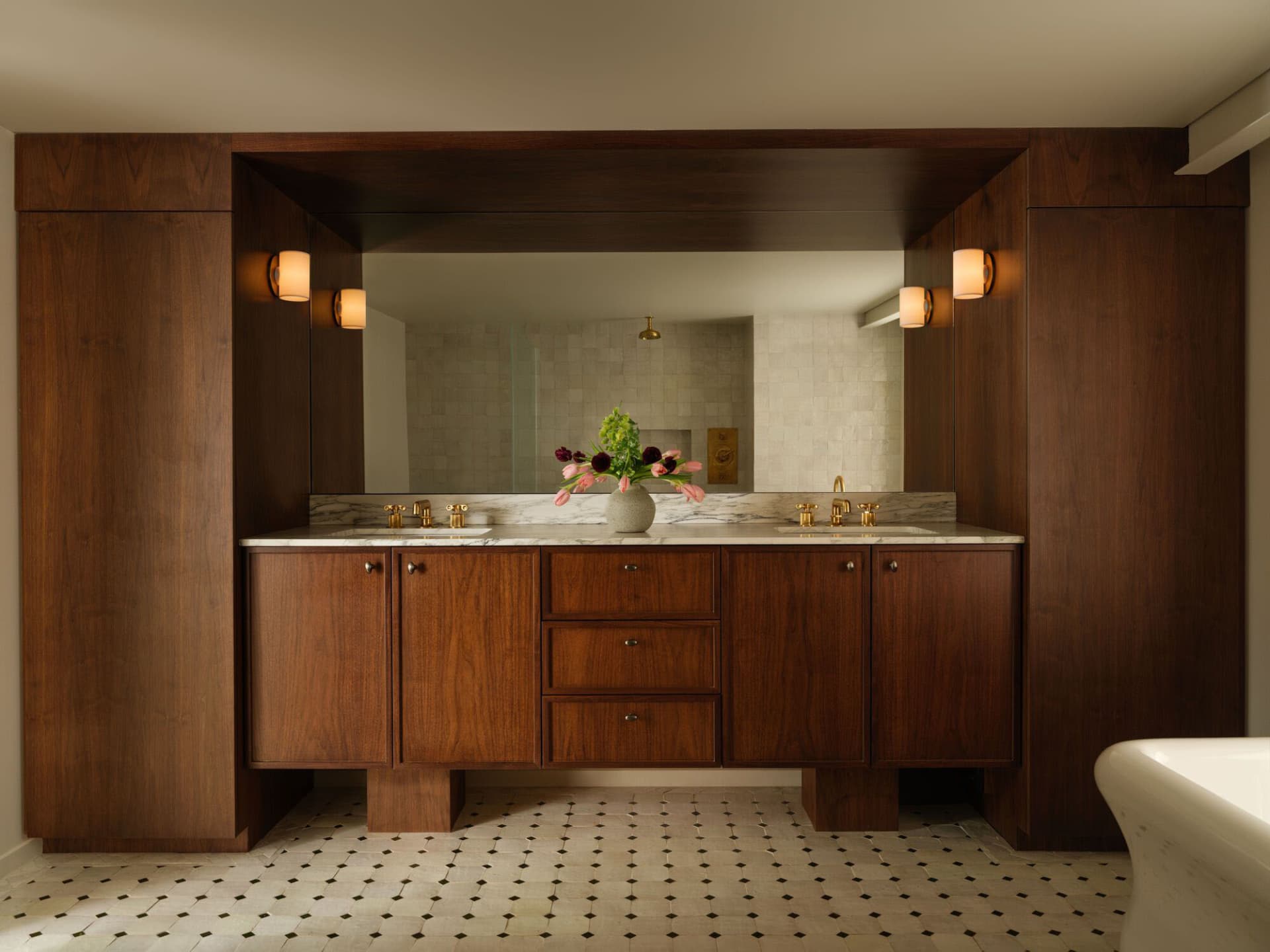 Luxurious bathroom with a double walnut vanity, white marble countertop and floor tiles, large mirror, and brass wall sconces.