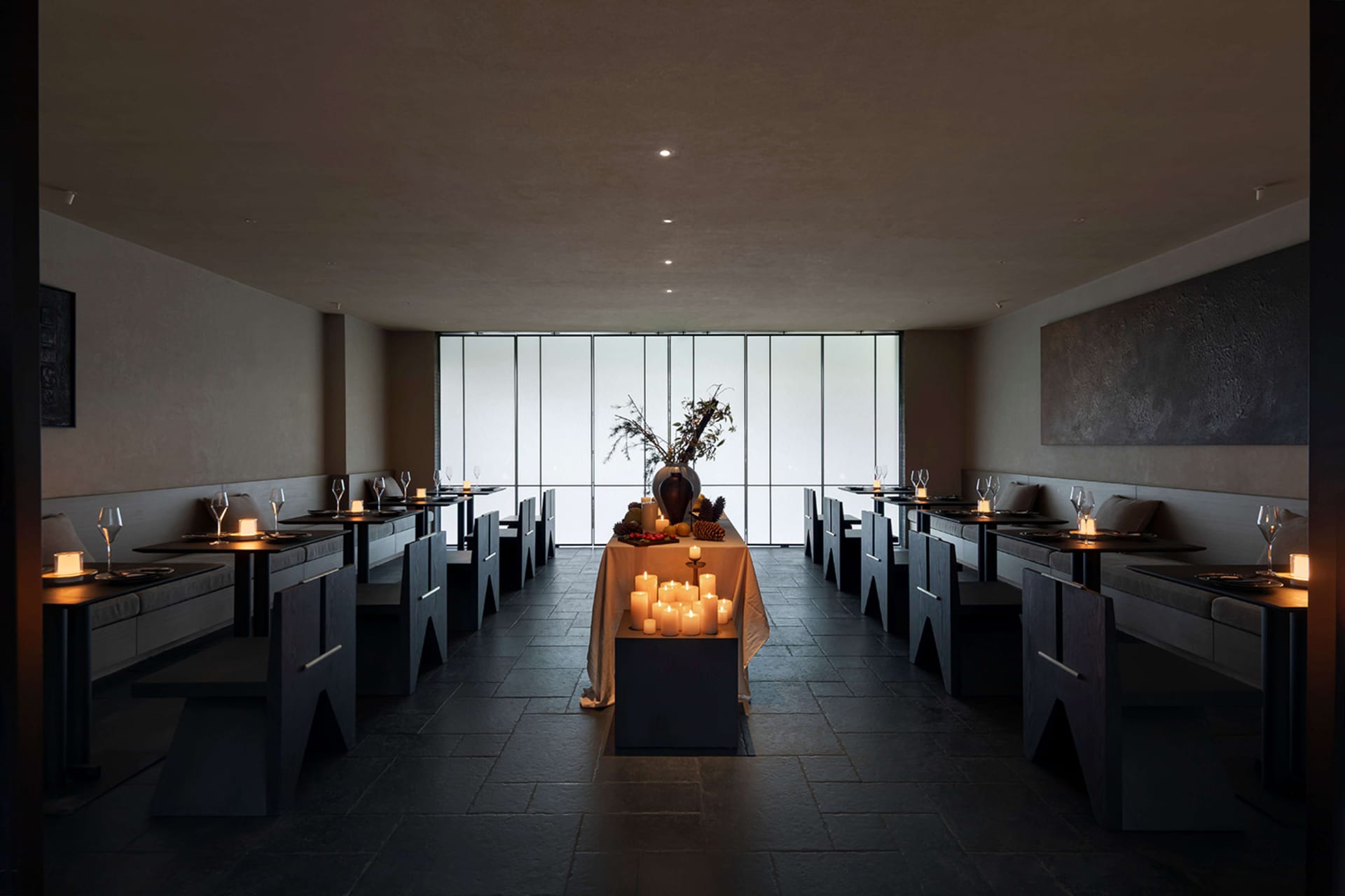 Interior view of a minimalist fine dining restaurant featuring dark stone floors and walls, built-in banquette seating, angular dark wood tables and chairs, and numerous lit candles creating warm ambient lighting, with large floor-to-ceiling windows at the far end.