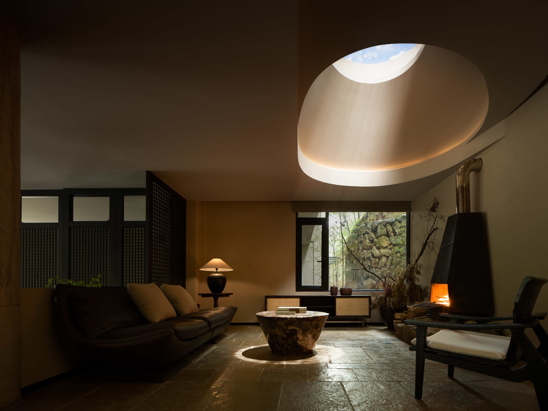 Modern minimalist living room featuring a large oval skylight, dark leather sofa, stone coffee table, black fireplace, and slate floor with natural light and warm tones.