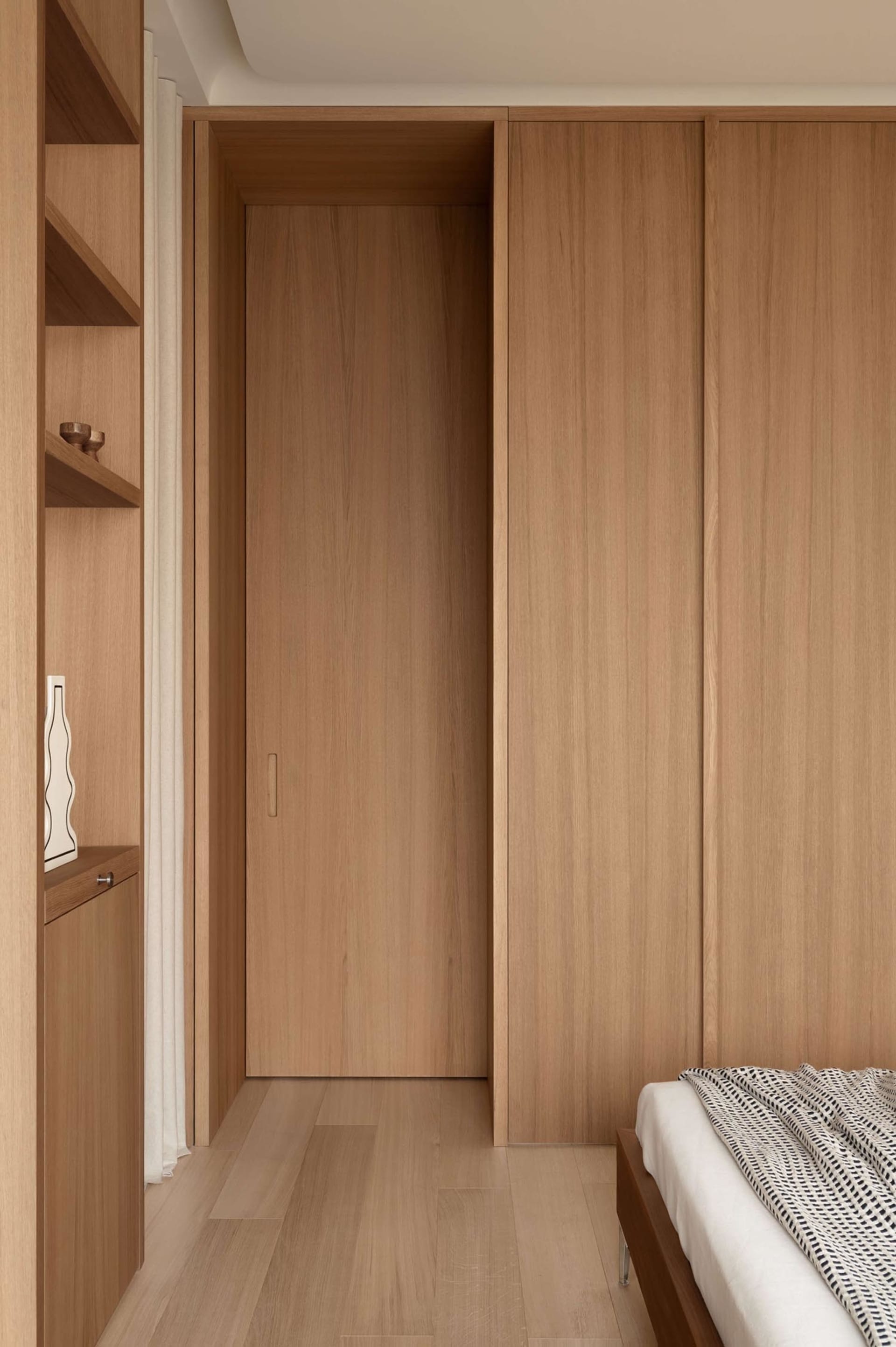 Modern minimalist bedroom featuring floor-to-ceiling light oak built-in wardrobe and open shelving, with a low wooden bed frame partially visible.