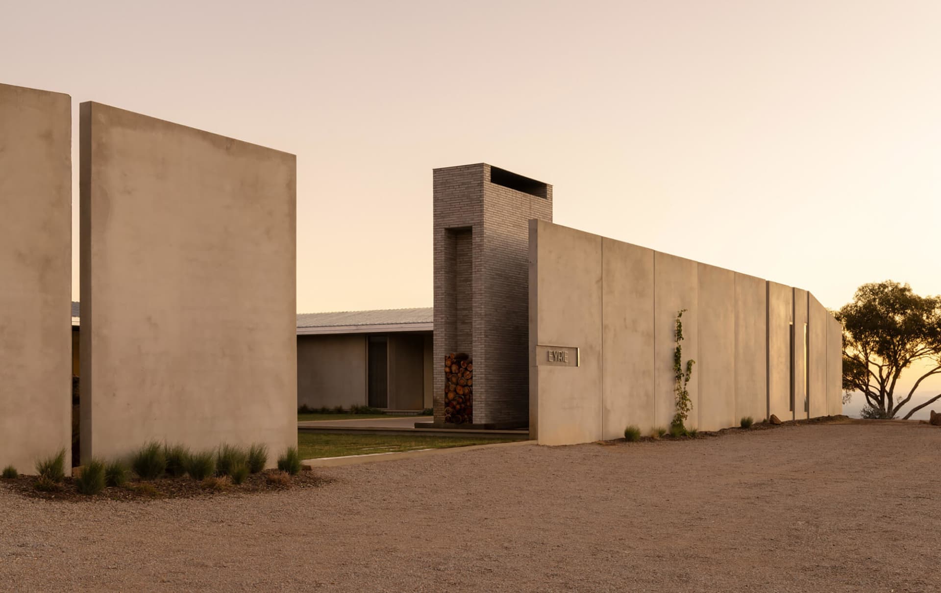 Modern concrete house named Eyrie with tall chimney, stacked firewood, and long horizontal walls at sunset in a rural setting.