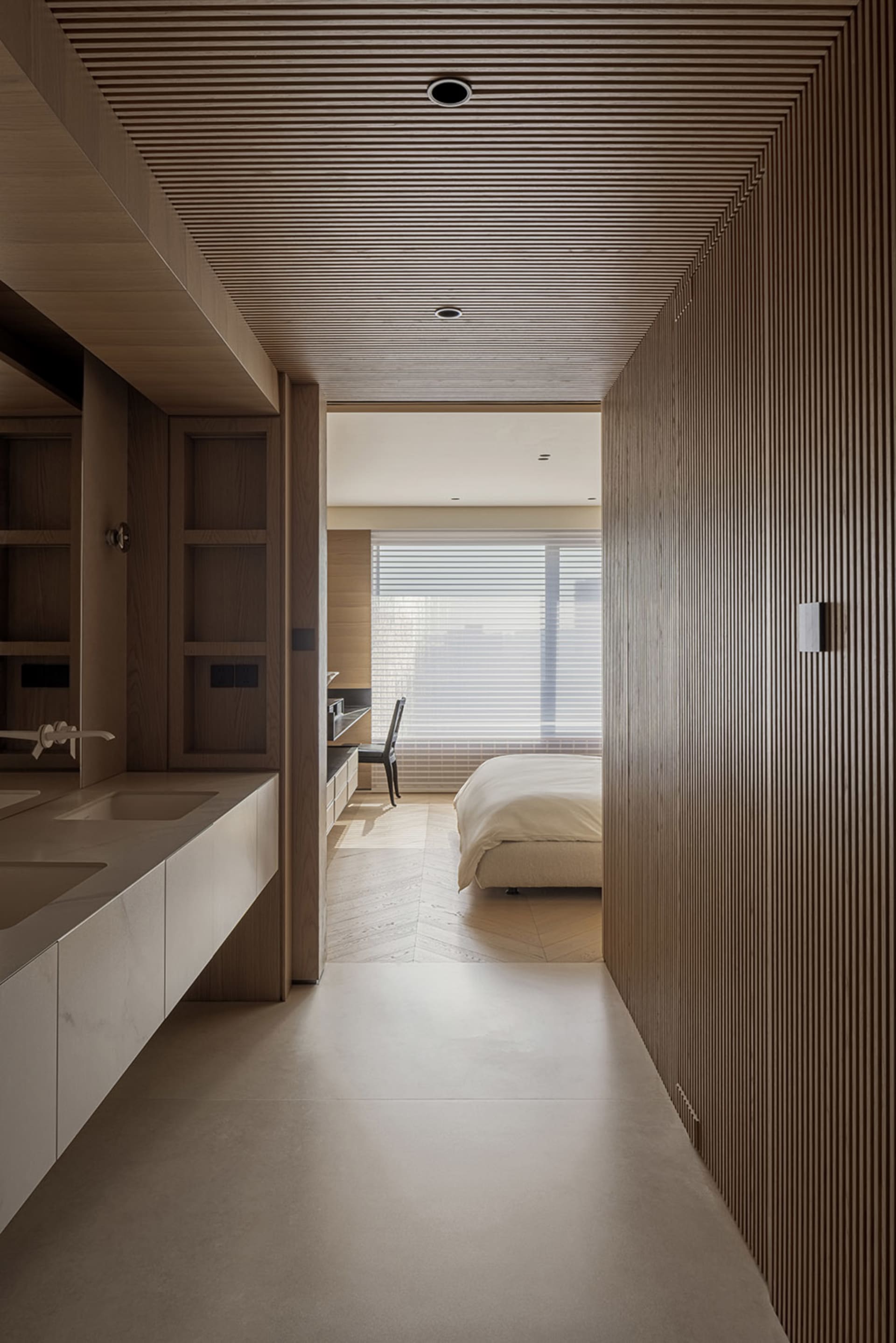 Modern minimalist bedroom viewed from an ensuite bathroom area featuring a marble double vanity, light wood cabinetry, vertical wood slat walls, and a bed with white linens in the background.