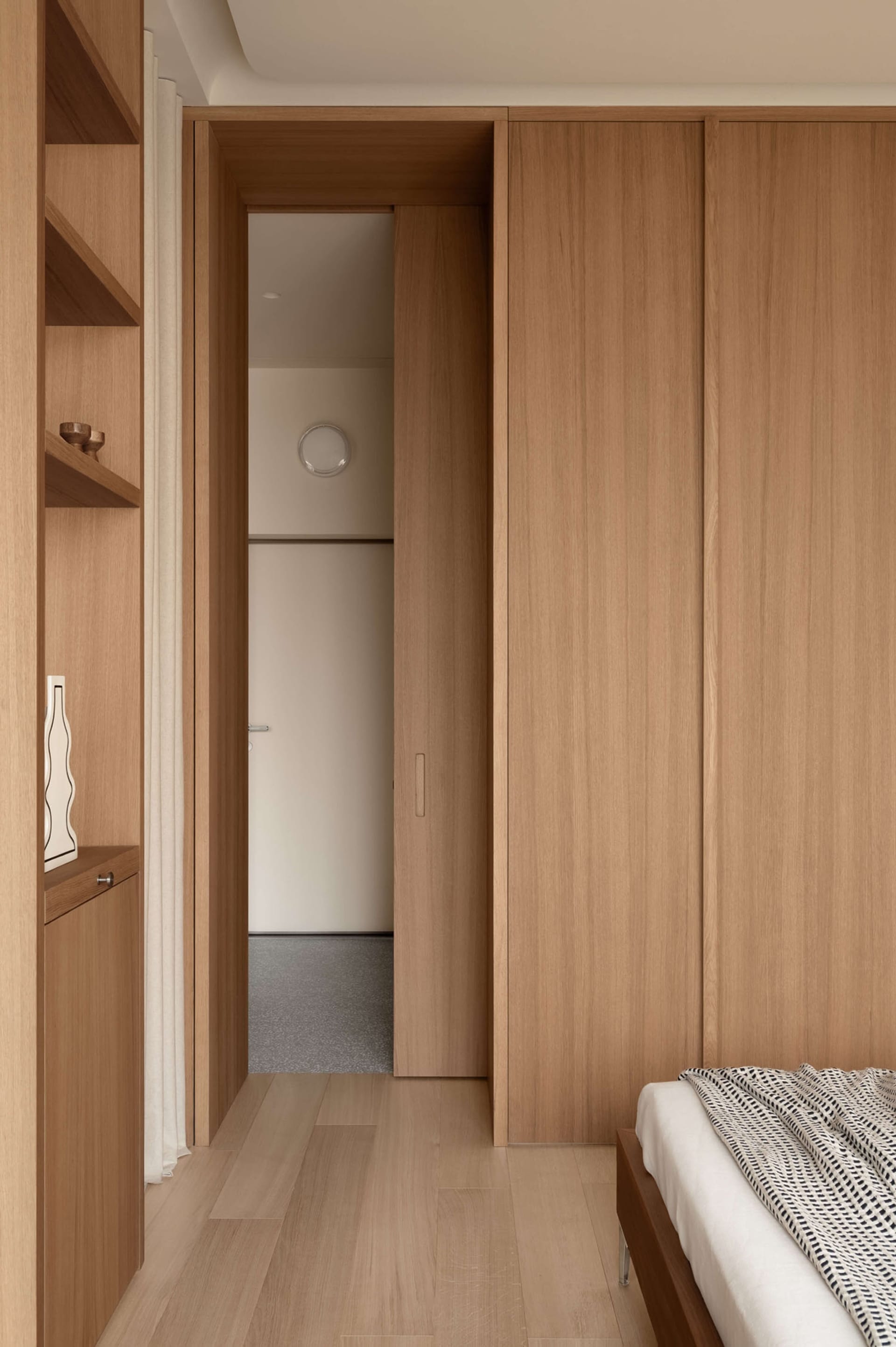 Modern minimalist bedroom interior with light oak built-in wardrobe, open wooden shelving, white bedding with black geometric throw, and a view through an open doorway to another room.