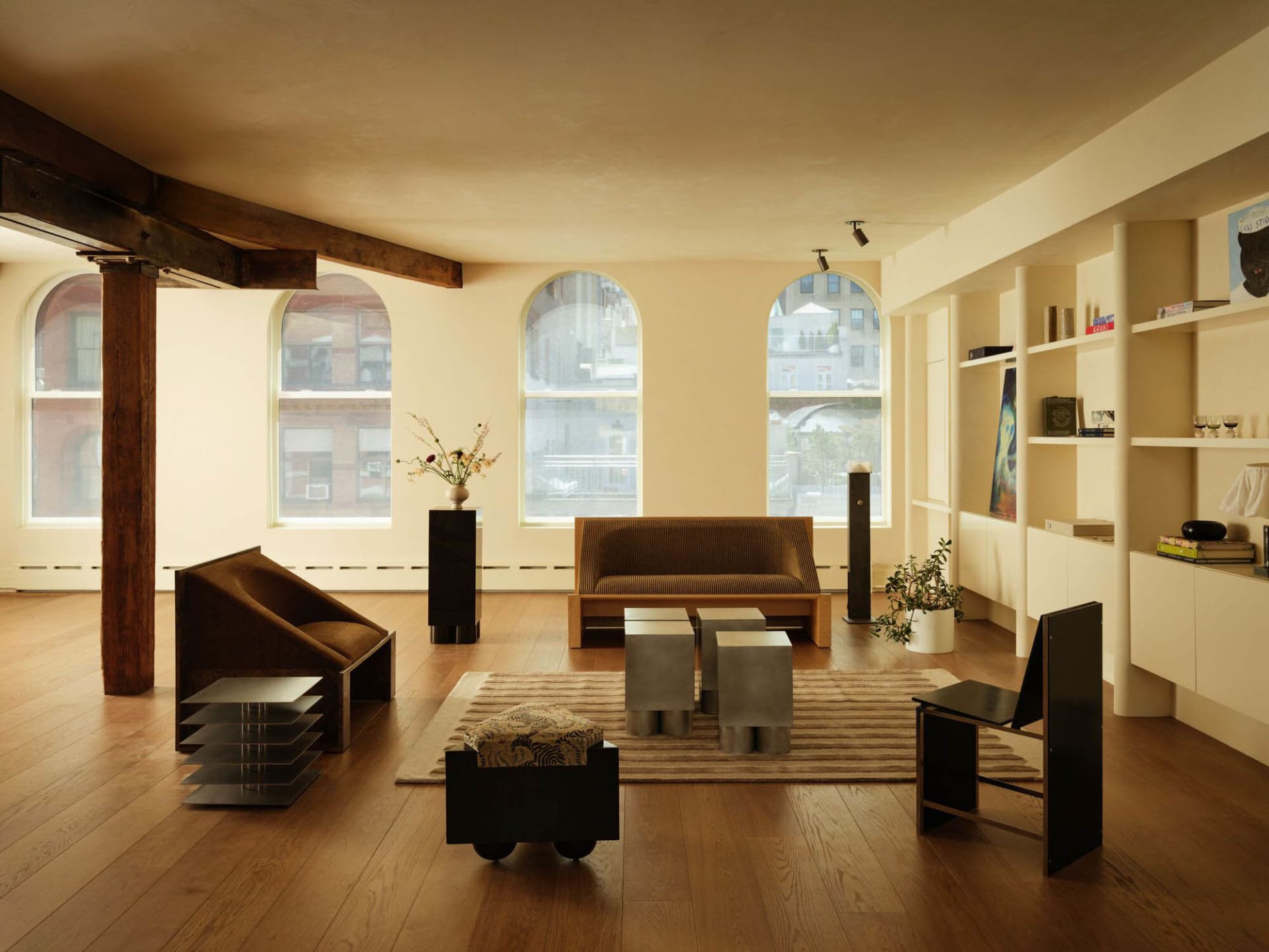 Spacious modern loft living room with dark walnut beams, arched windows, corduroy sofa, sculptural brown armchair, concrete side tables, and built-in shelving.