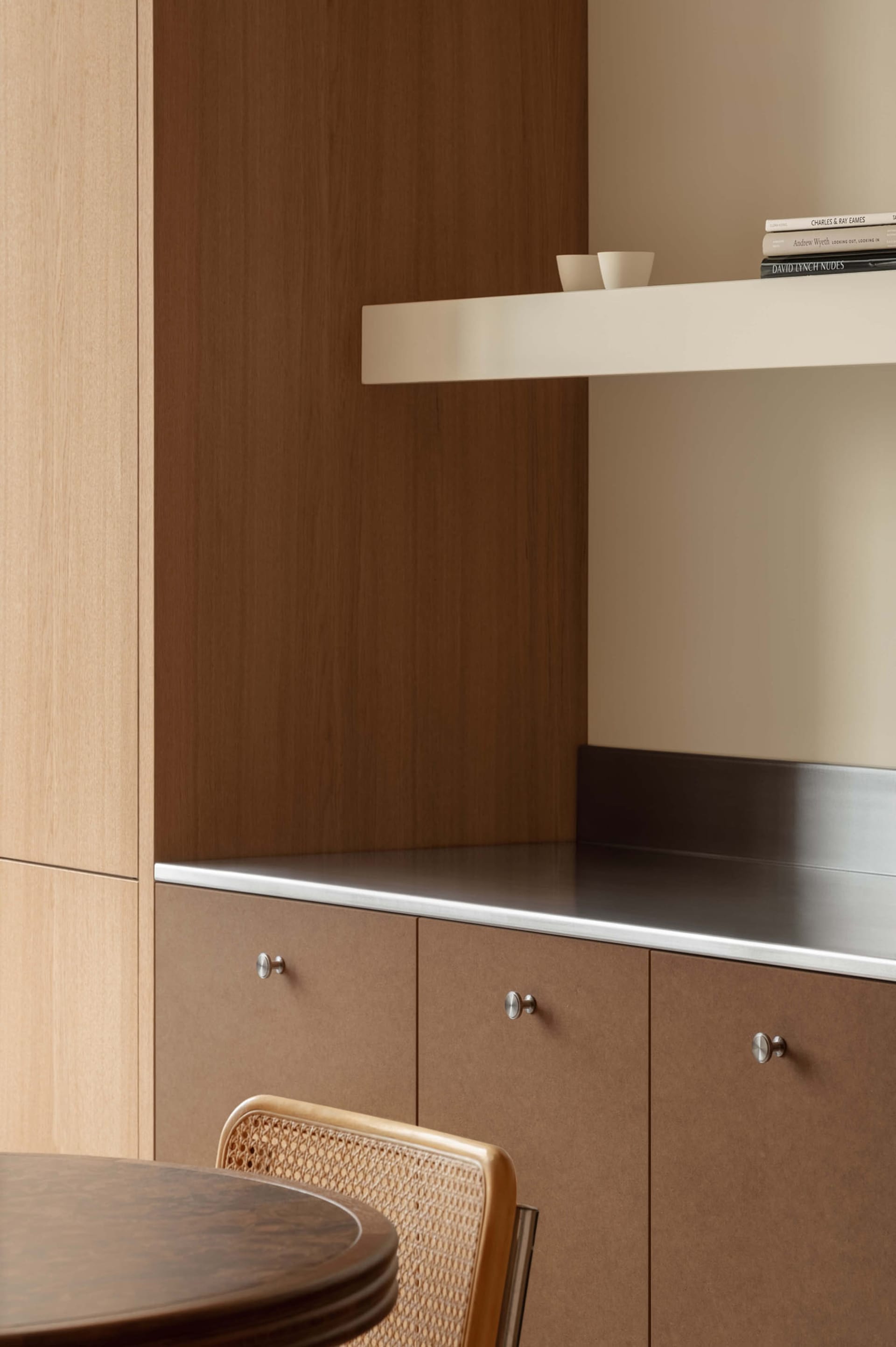 Close-up of a modern dining area featuring warm walnut lower cabinets with round silver knobs, a stainless steel countertop, light oak upper cabinetry, and a cane-backed dining chair in the foreground.