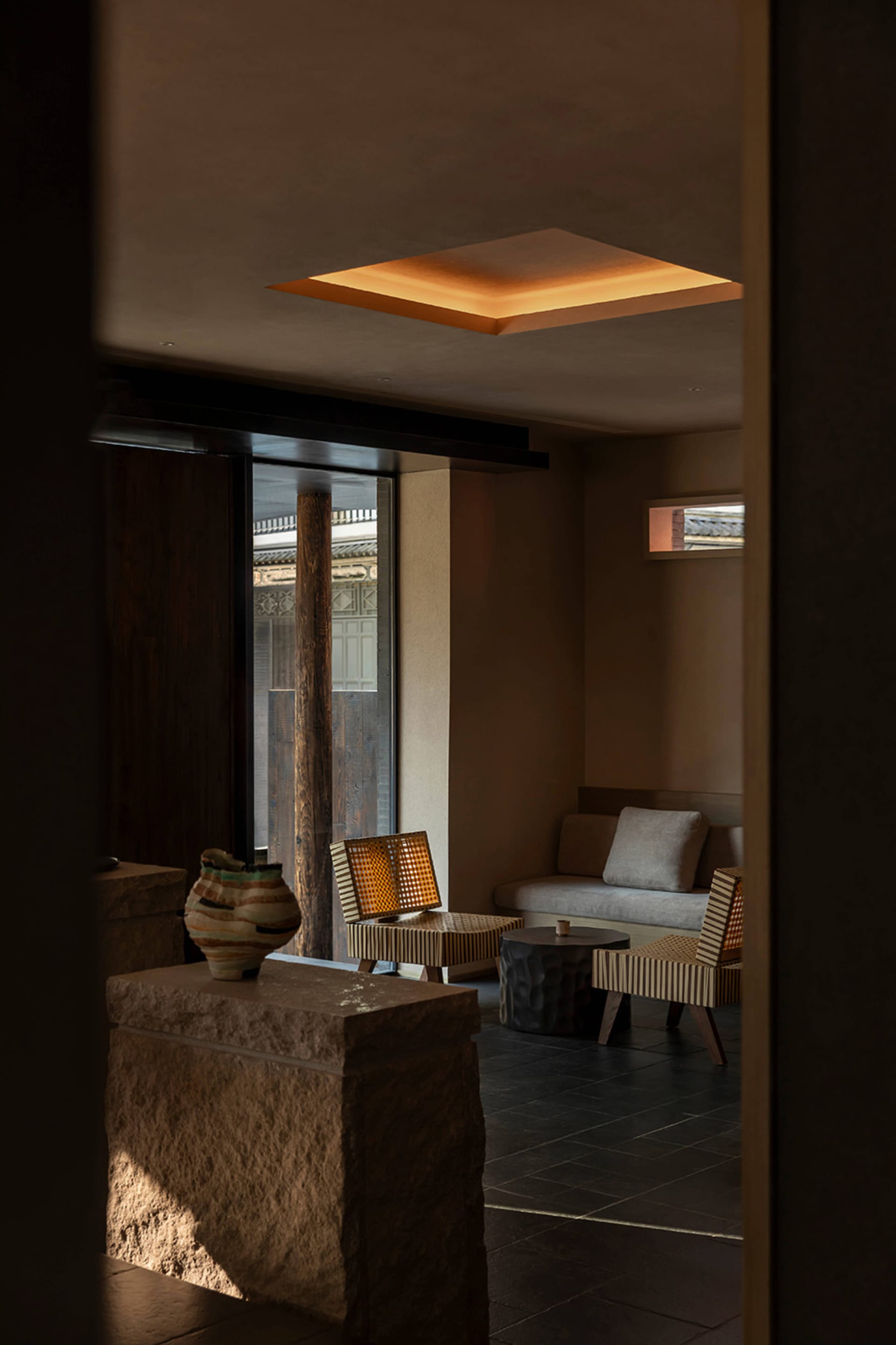 Modern minimalist lounge with raw stone blocks, woven rattan chairs, dark tiled floor, and a glowing square skylight in an earthy-toned interior.
