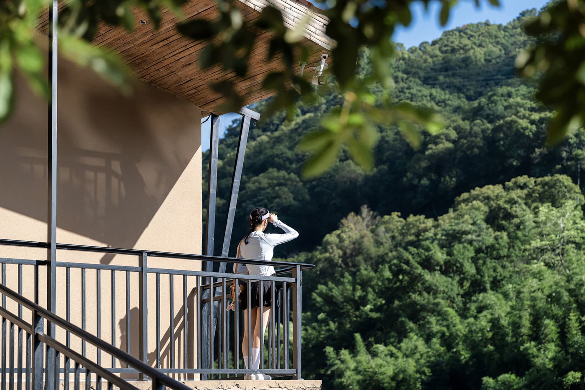 Woman in athletic wear standing on a modern balcony with wooden ceiling and metal railing, looking at a forested mountain landscape.