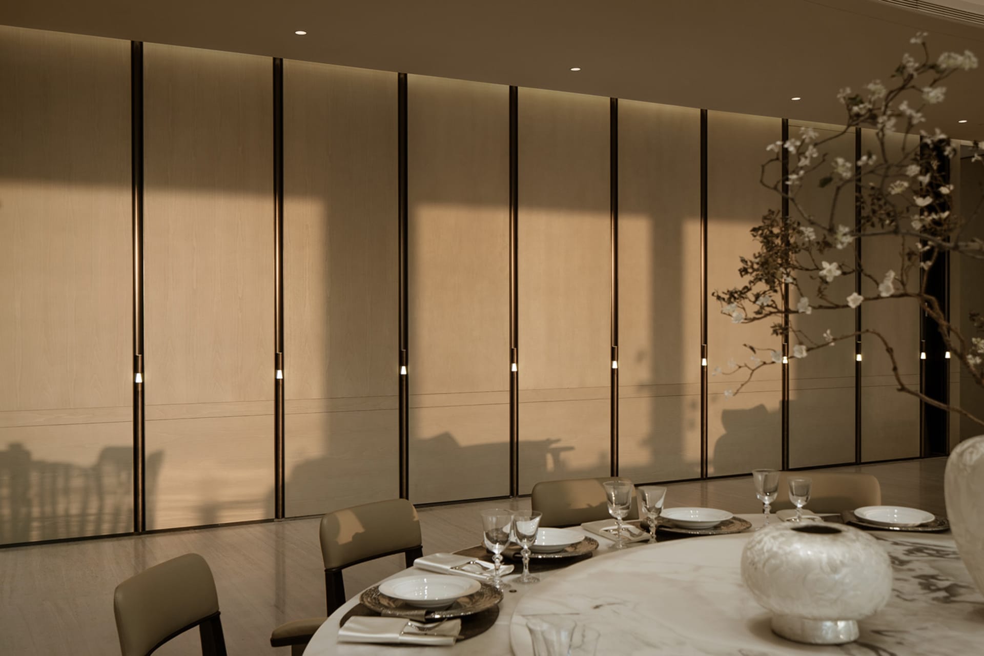 Elegant modern dining room with a round white marble table set for dinner, taupe leather chairs, and a dramatic floor-to-ceiling backlit oak panel wall with vertical light accents.