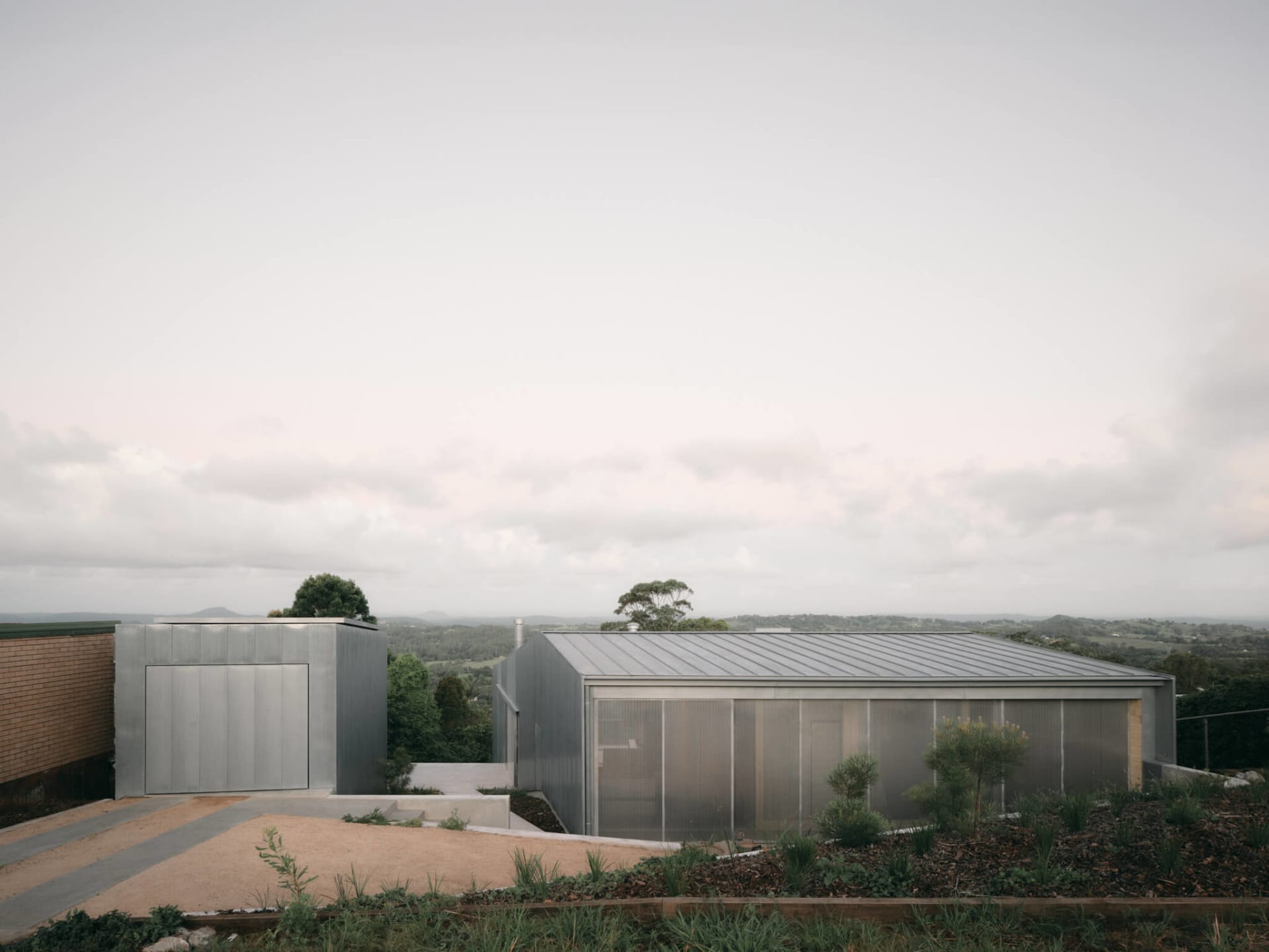 Modern single-story house with corrugated metal cladding and large translucent windows, situated on a hilltop overlooking a rural valley landscape under an overcast sky.