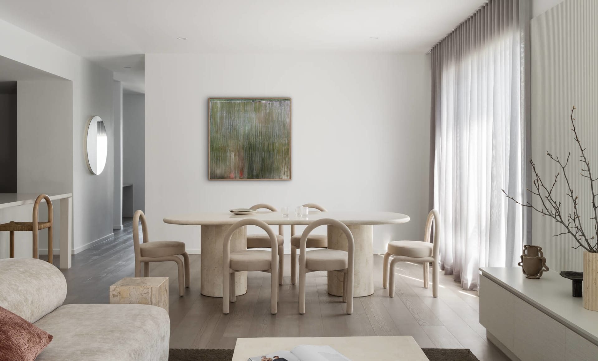 Modern minimalist dining room featuring a large travertine oval table, six curved upholstered chairs, abstract green painting, and sheer curtains allowing natural light.