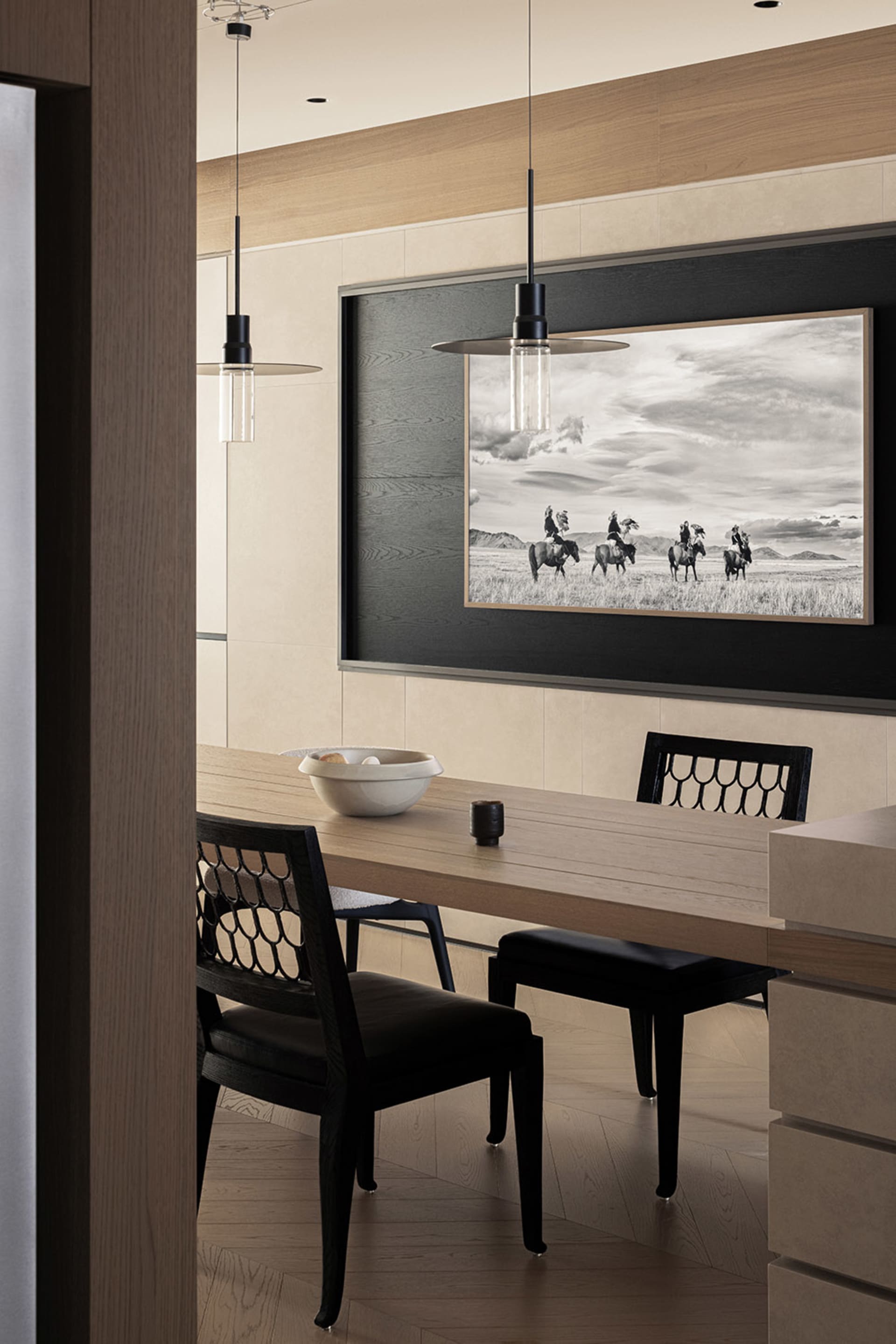 Modern dining room featuring a light oak table, black lattice-back chairs, two black pendant lights, and a large black-and-white photograph of cowboys on horseback.
