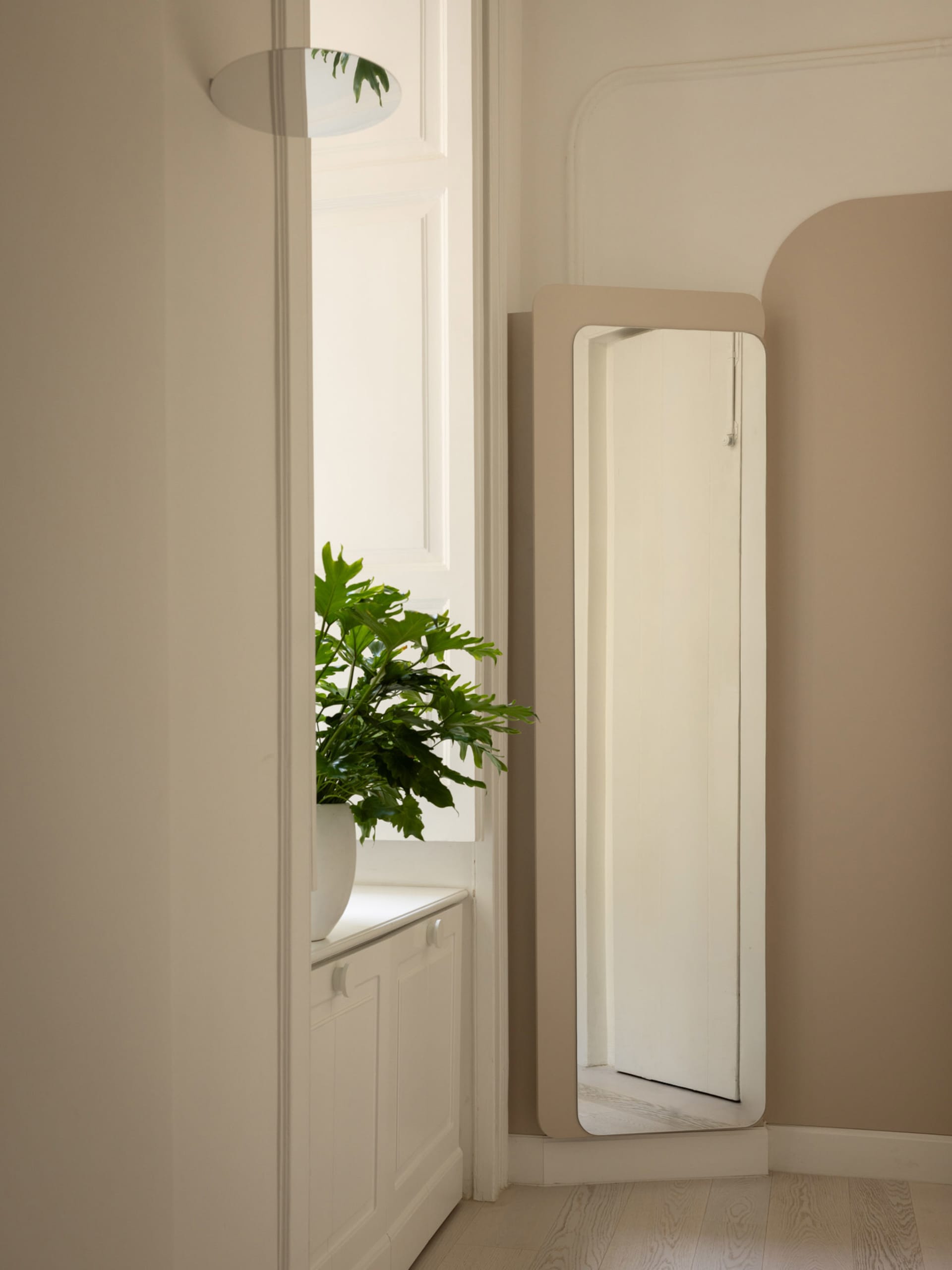 Minimalist entryway corner featuring a tall full-length mirror with a matte beige frame, a white arched wall detail, and a large potted monstera plant on a white cabinet by the window.
