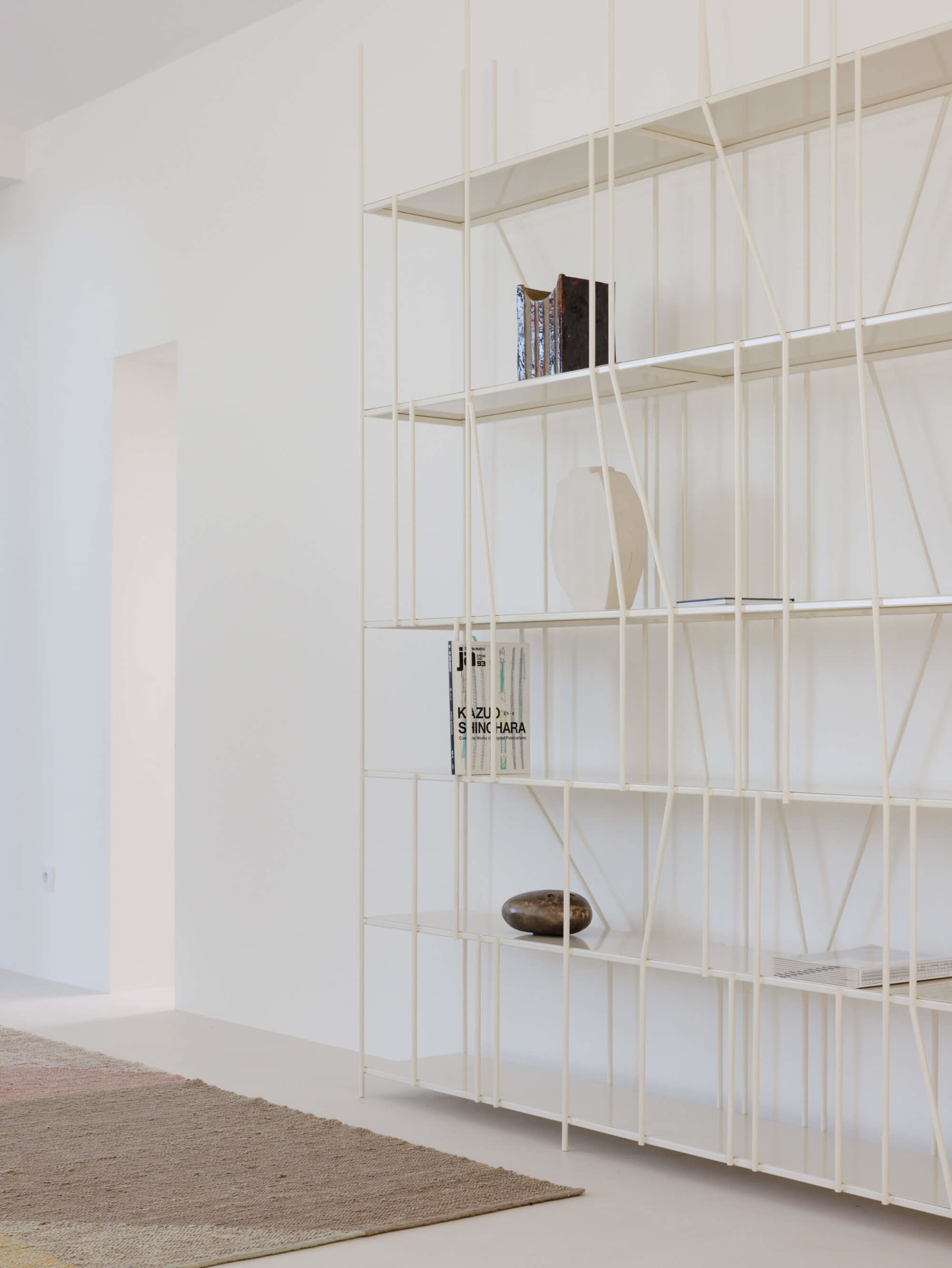 Modern white metal grid bookshelf against a white wall, displaying books, ceramic vases, and sculptural objects, with a textured area rug in the foreground.