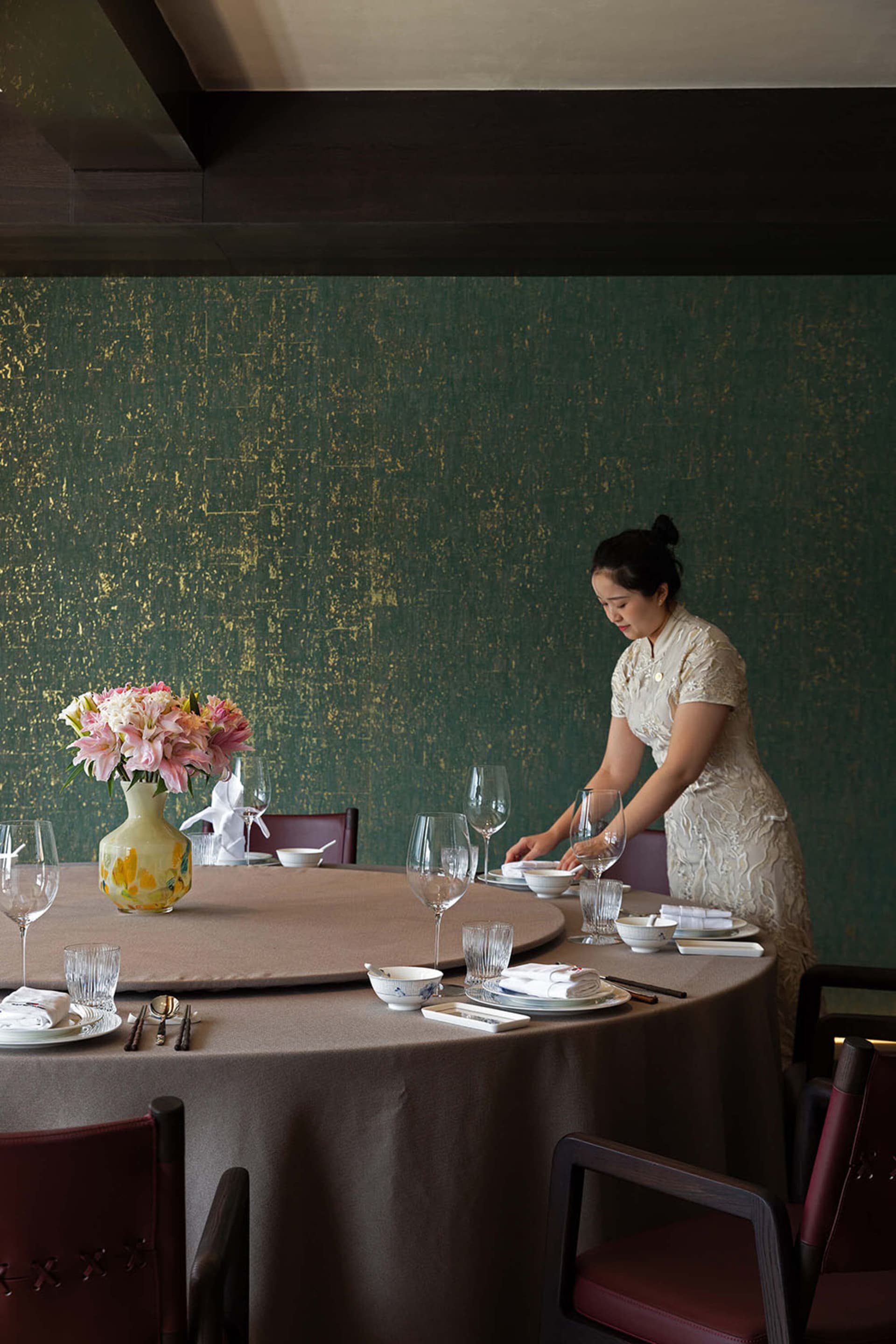Elegant Chinese fine dining room featuring a large round table set with porcelain and crystal, set against a deep emerald green textured wall with gold leaf accents, with a server in a cream lace qipao arranging tableware.