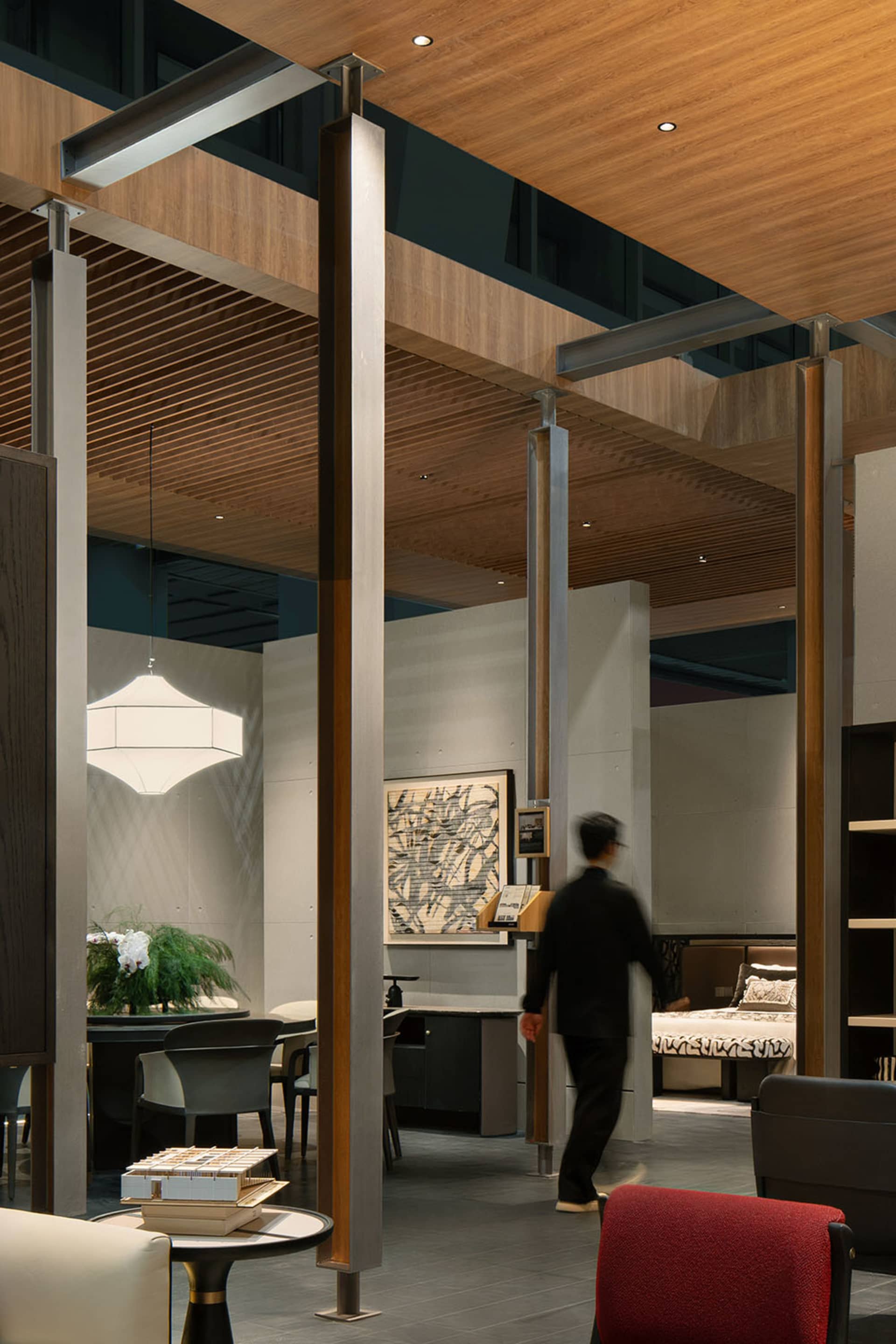 Modern commercial showroom interior with wooden ceiling slats, concrete structural columns, architectural model display, dining area, and bed vignette in the background.