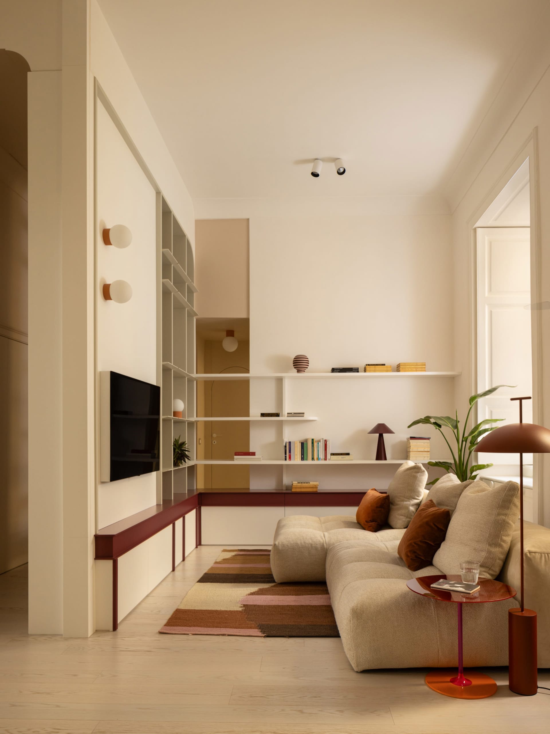 Modern minimalist living room with off-white walls, built-in white shelving, terracotta-red accents, a beige modular sofa with brown and beige cushions, a striped area rug, and a large potted plant.